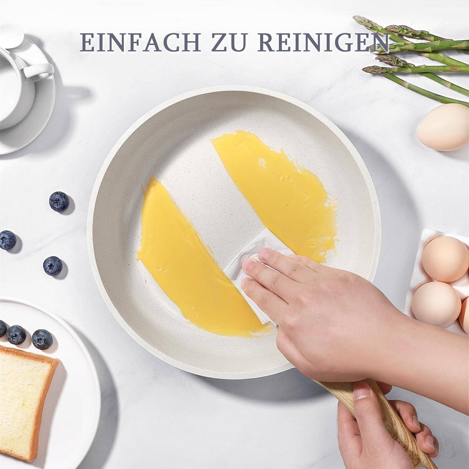 Eine Hand wischt eine Pfanne mit einem gelben Schwamm ab. Blaubeeren, Brot, Eier, Spargel und eine Tasse befinden sich in der Nähe. Text lautet: 