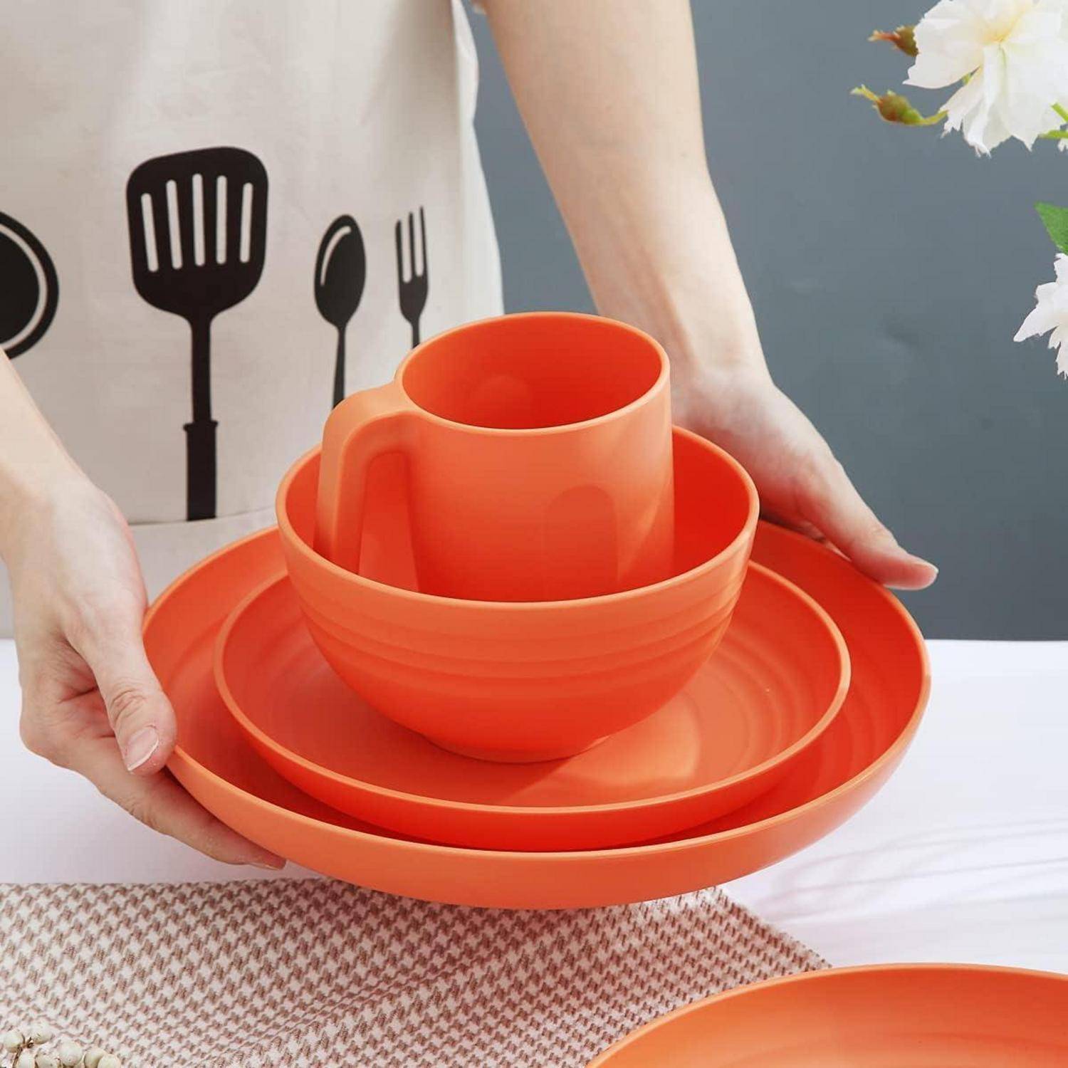 Eine Person ordnet einen Stapel orangefarbener Geschirrstücke an, darunter einen Teller, eine Schüssel und eine Tasse, auf einer Arbeitsplatte. Sie trägt eine Schürze mit Küchengerät-Designs.