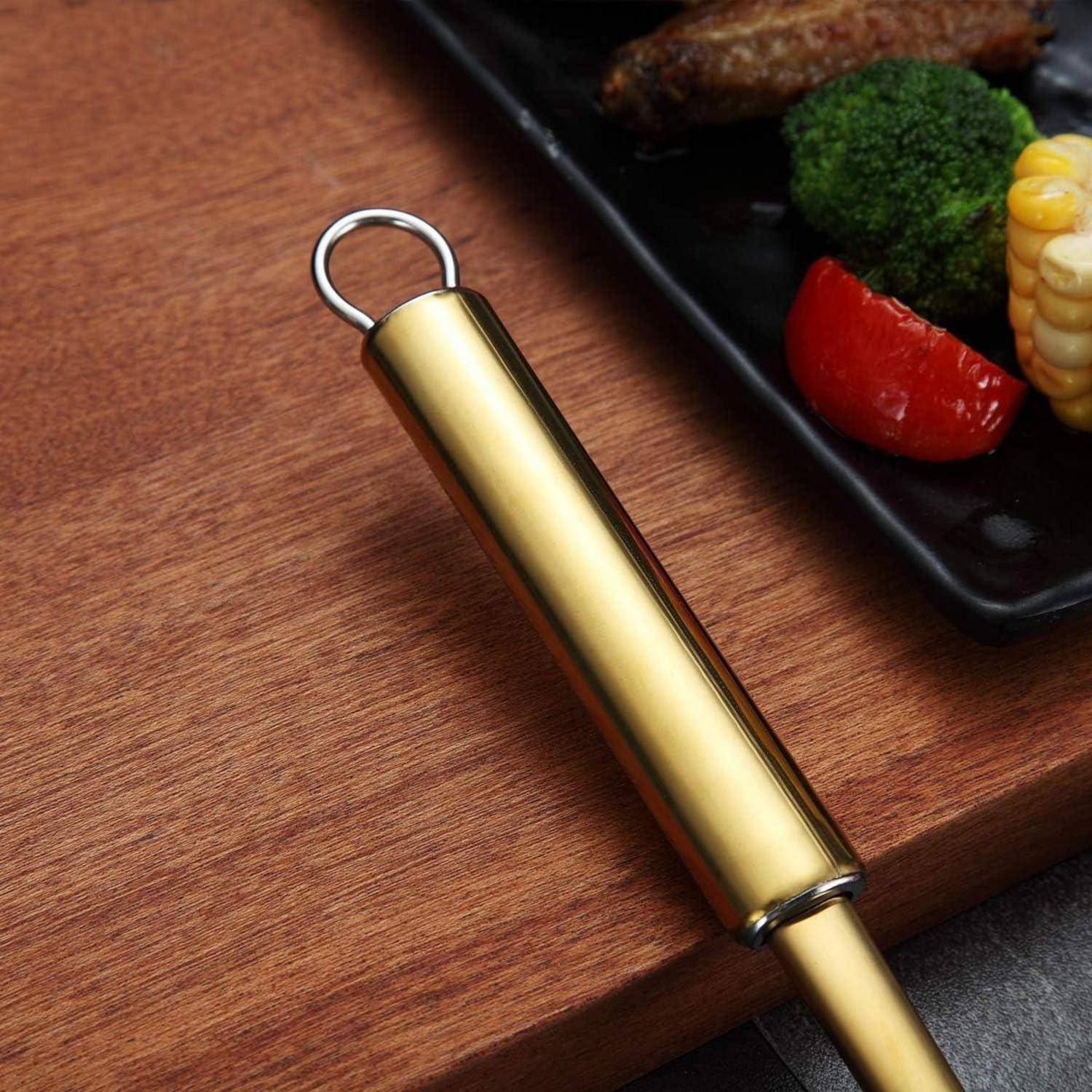 Ein goldener Griff eines Küchenutensils liegt auf einer Holzoberfläche, neben einem schwarzen Teller mit Brokkoli, einem Fleischstück und einem Stück roter Paprika.