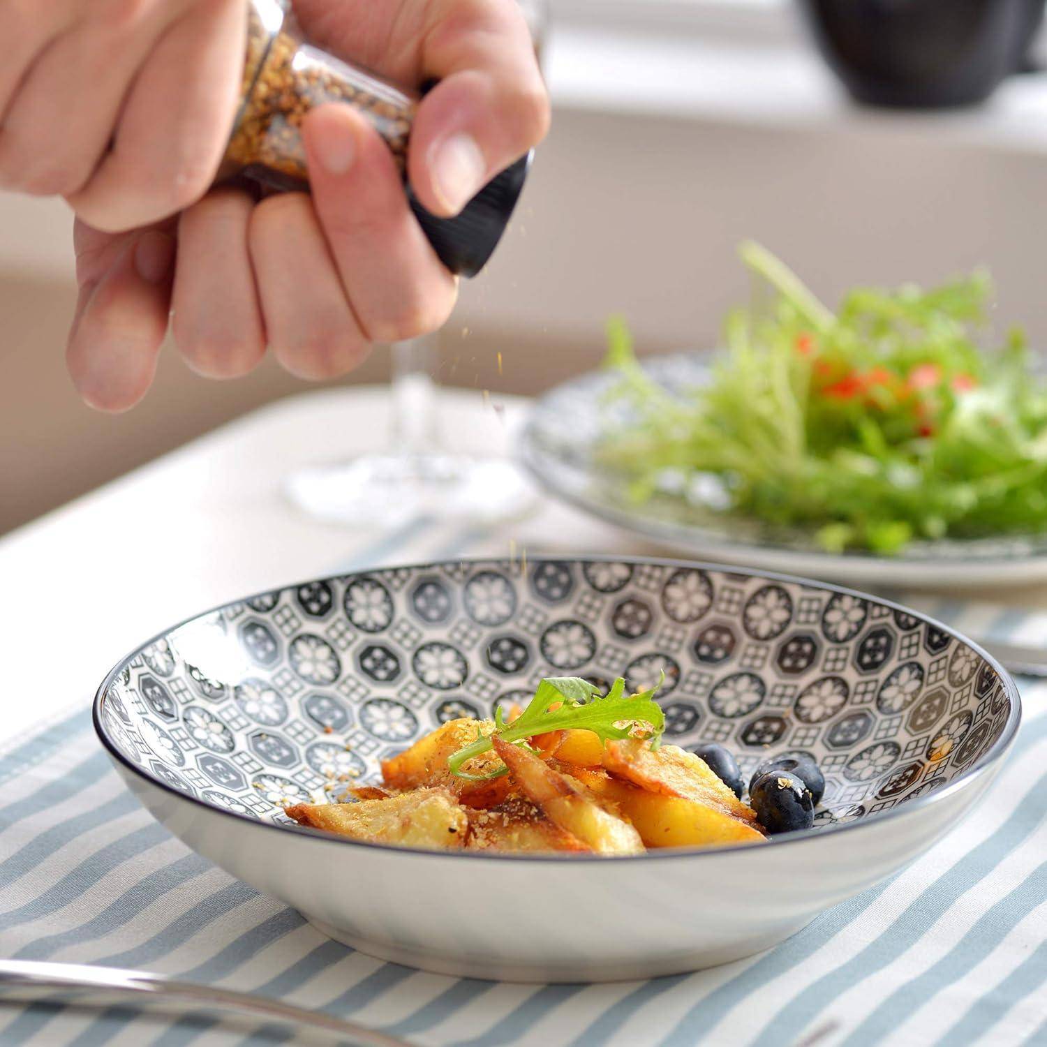 Eine Hand streut Gewürze über eine dekorative Schüssel Gnocchi, die mit Kräutern garniert ist. Ein Salat steht im Hintergrund auf einem Tisch.