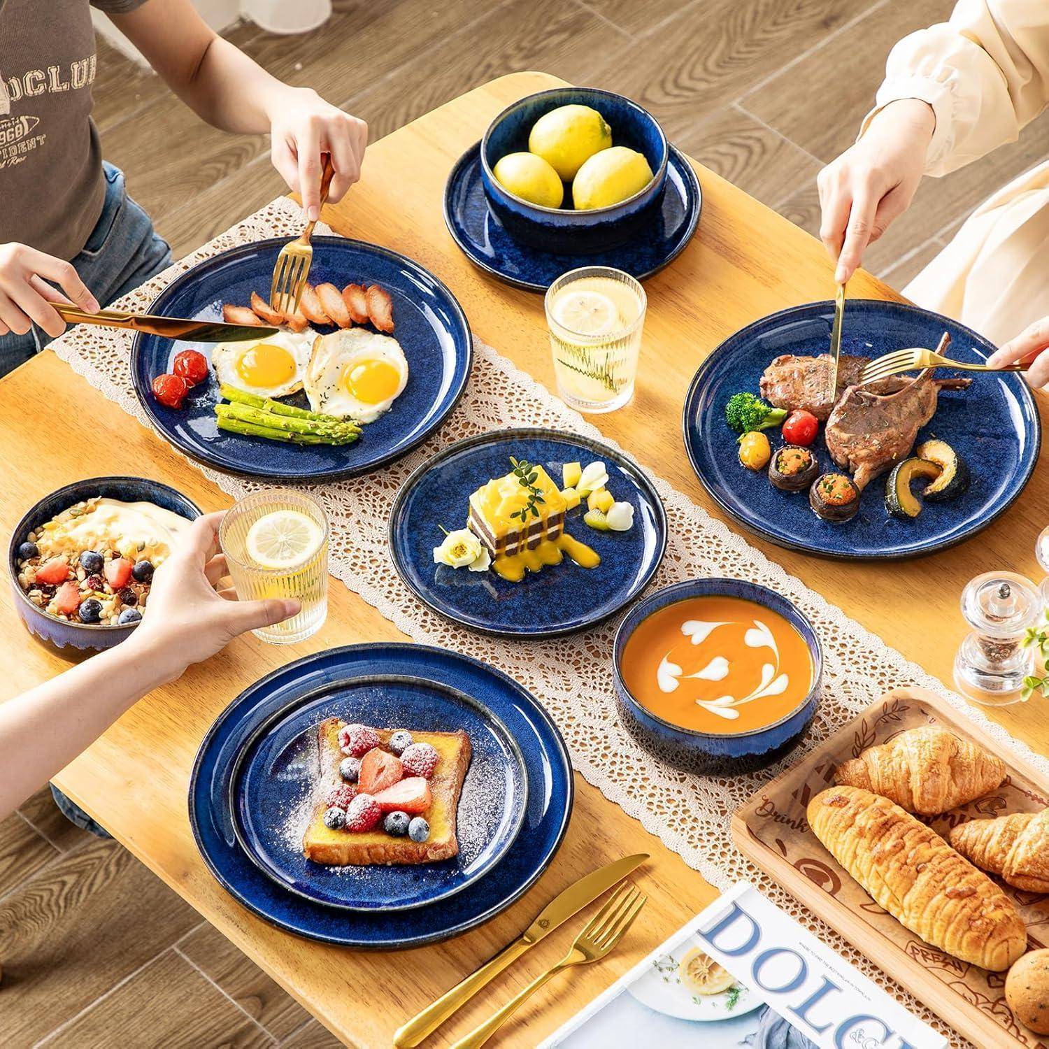 Ein Esstisch mit verschiedenen Gerichten, darunter Steak, Eier mit Spargel, Suppe, Pfannkuchen mit Beeren, Obstsalat und einem Korb mit Croissants. Menschen greifen nach Essen und Getränken.