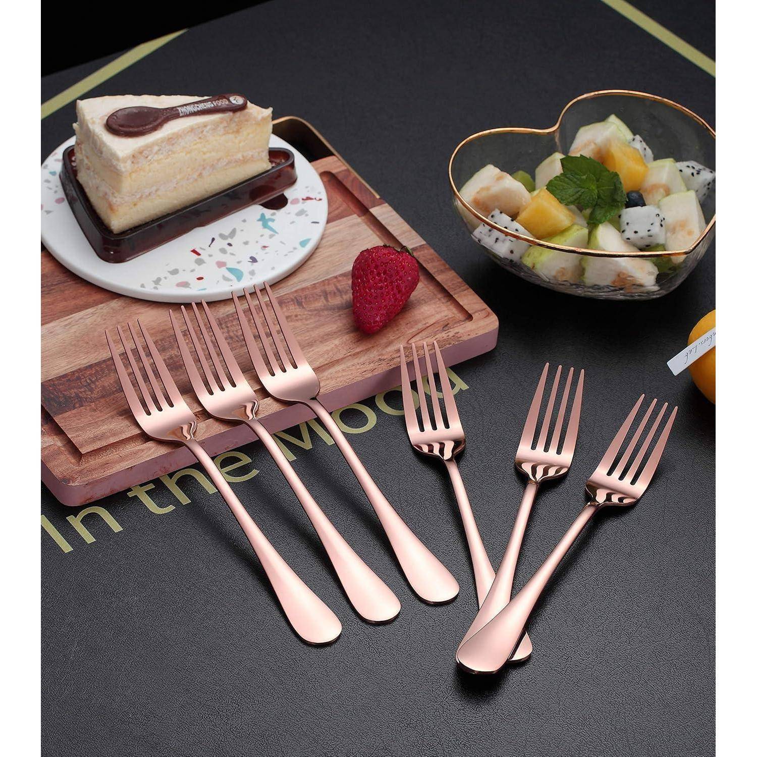 Eine Anordnung von sechs Roségold-Gabeln auf einer dunklen Oberfläche neben einem Holzbrett mit einem Kuchenstück und einer Schüssel mit gemischten Früchten, was einen Essensbereich andeutet.