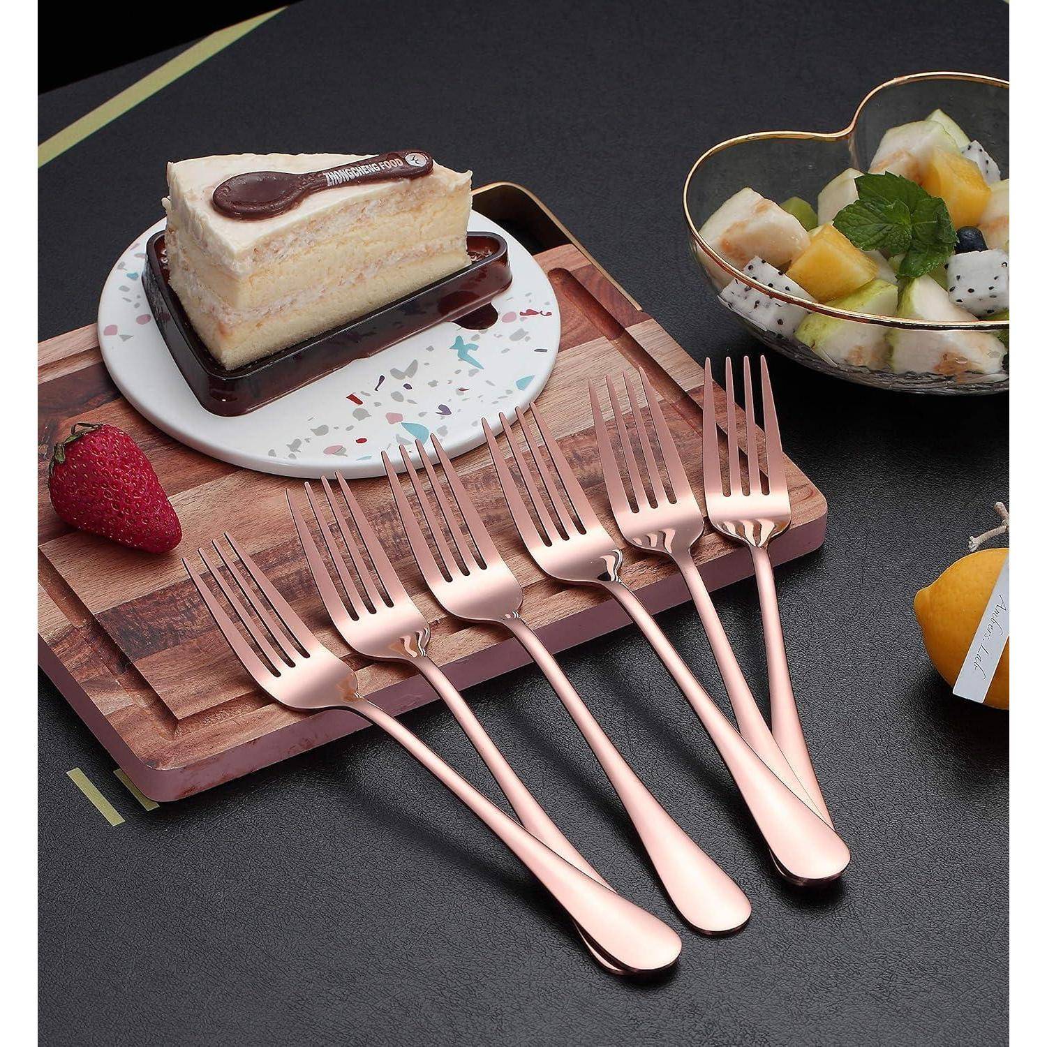 Ein Holztablett trägt eine Kuchenscheibe mit einem dekorativen Schokoladenlöffel und einer Erdbeere. Sechs roségoldene Gabeln sind davor angeordnet. Eine herzförmige Schüssel mit Obst befindet sich neben dem Tablett.