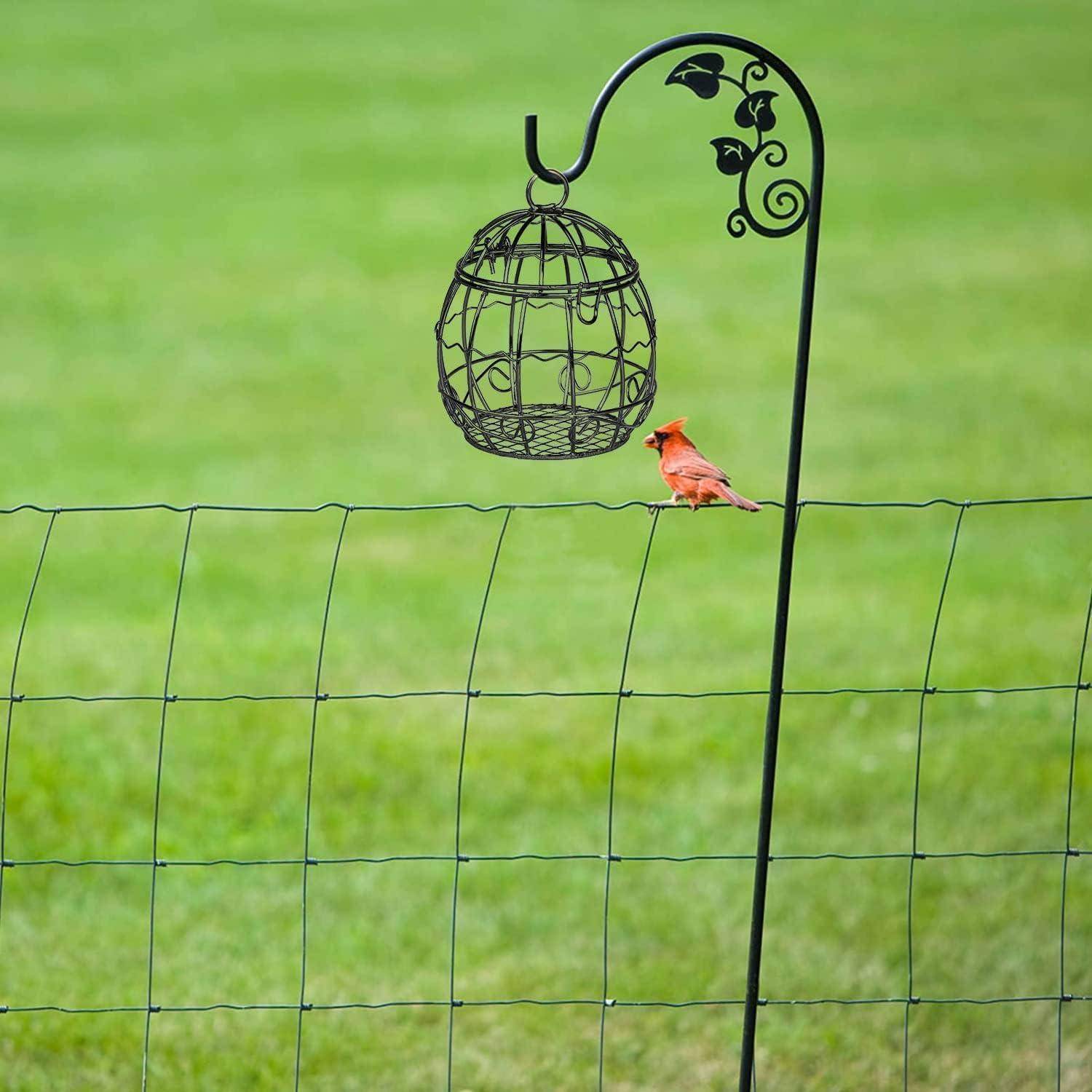 Ein roter Vogel sitzt auf einem Drahtzaun unter einem dekorativen hängenden Vogelkäfig, der sich in einem Grasfeld befindet.
