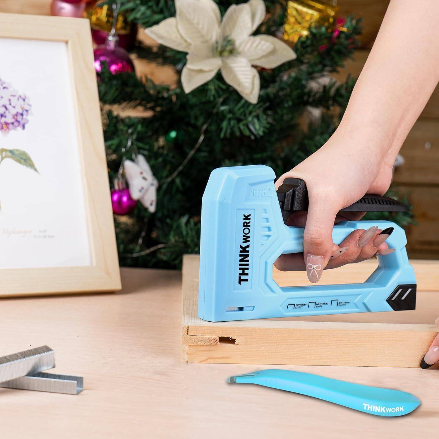 Eine Person, die eine blaue Tacker an einem Holzbrett in der Nähe eines Weihnachtsbaums verwendet, mit Dekorationen und einem Blumenkunstwerk im Hintergrund.