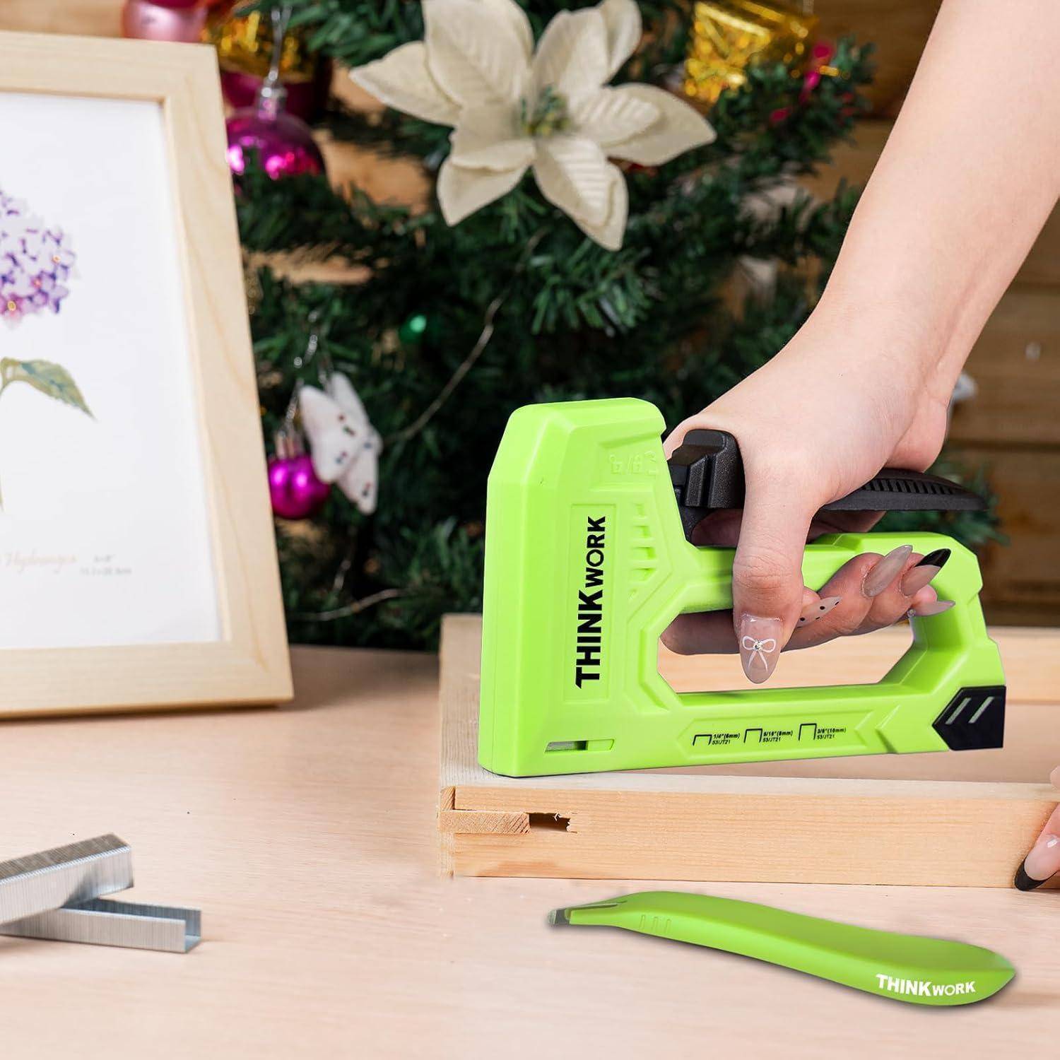 Eine Person verwendet eine grüne 'THINK WORK' Tacker-Pistole auf einer Holzoberfläche. Ein Weihnachtsbaum und gerahmte Blumenkunst befinden sich im Hintergrund.
