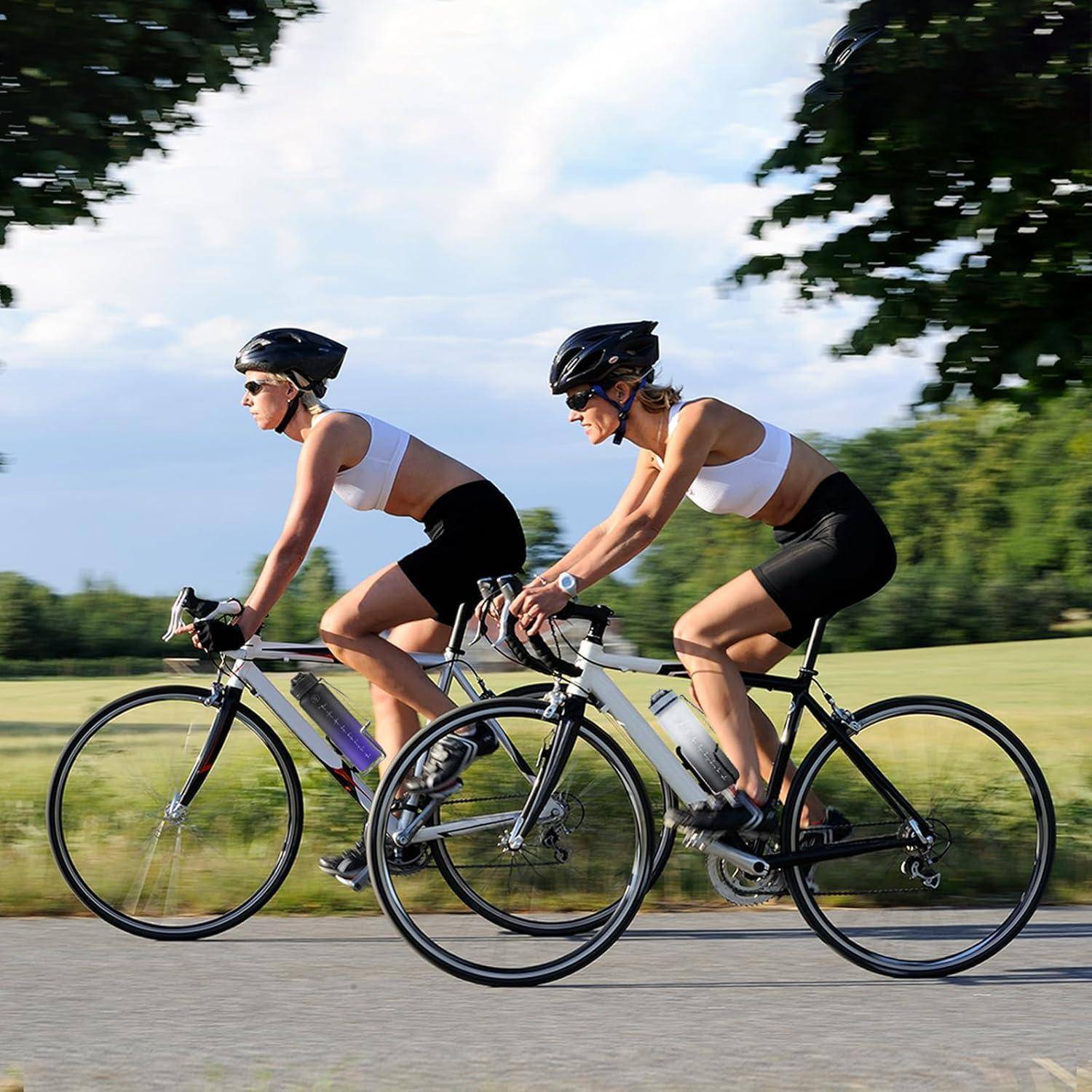 Zwei Personen mit Helmen und Sportkleidung fahren Fahrrad auf einer Landstraße mit Feldern und Bäumen im Hintergrund.