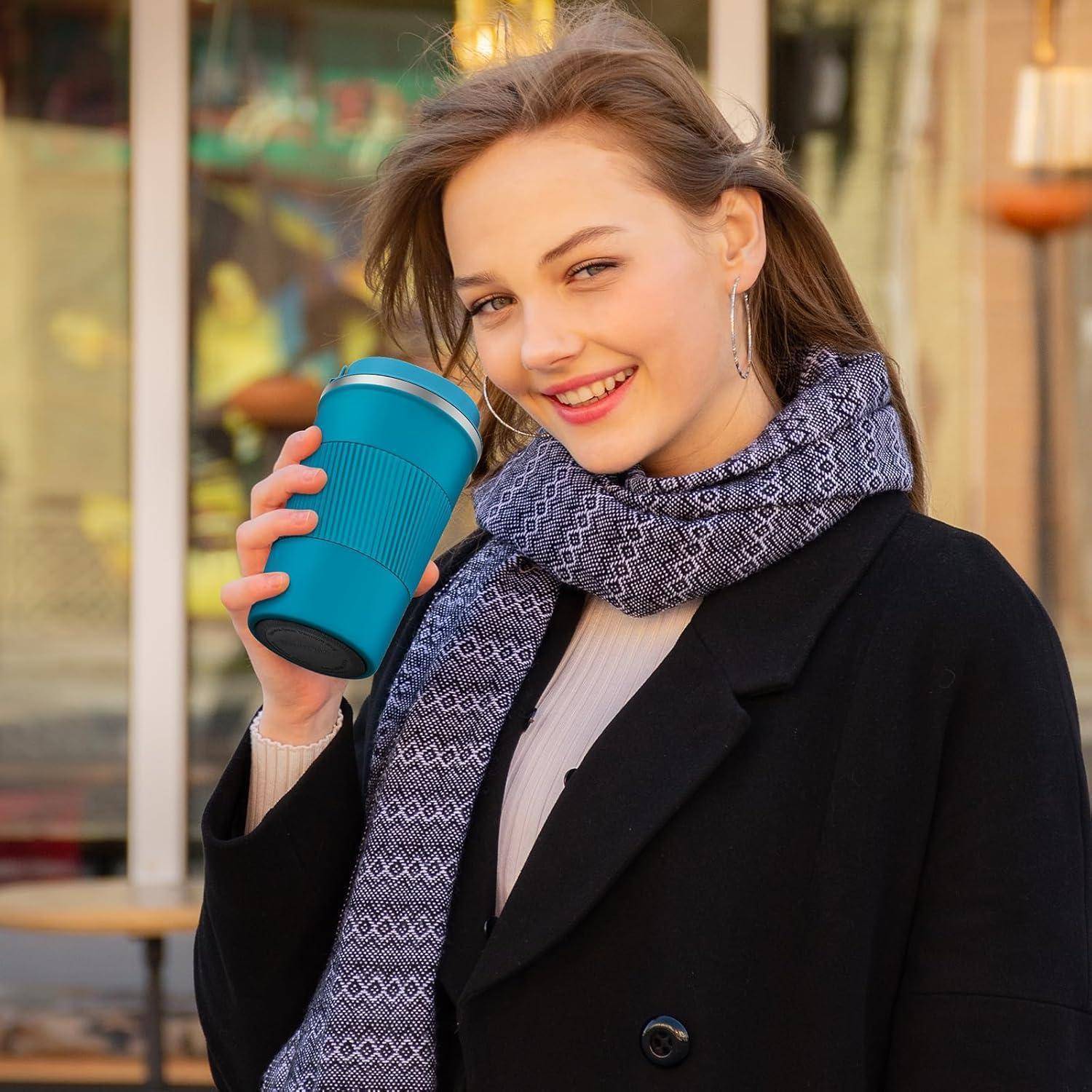 Eine lächelnde Person hält einen wiederverwendbaren Kaffeebecher und steht draußen, trägt einen gemusterten Schal und einen Mantel, was auf einen kühlen Herbsttag hindeutet.