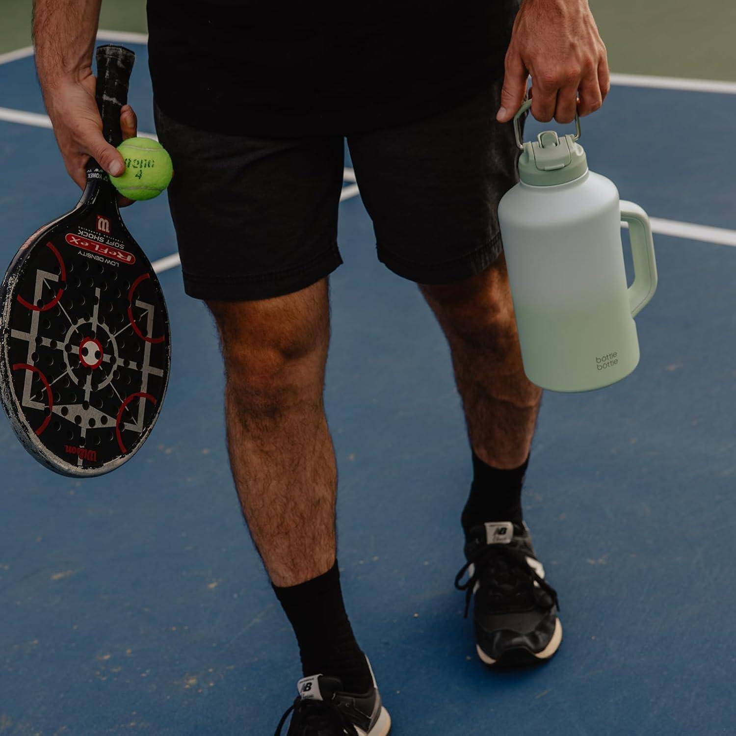 Eine Person, die einen Paddel, ein Pickleball und eine große Wasserflasche hält und auf einem Sportplatz steht.