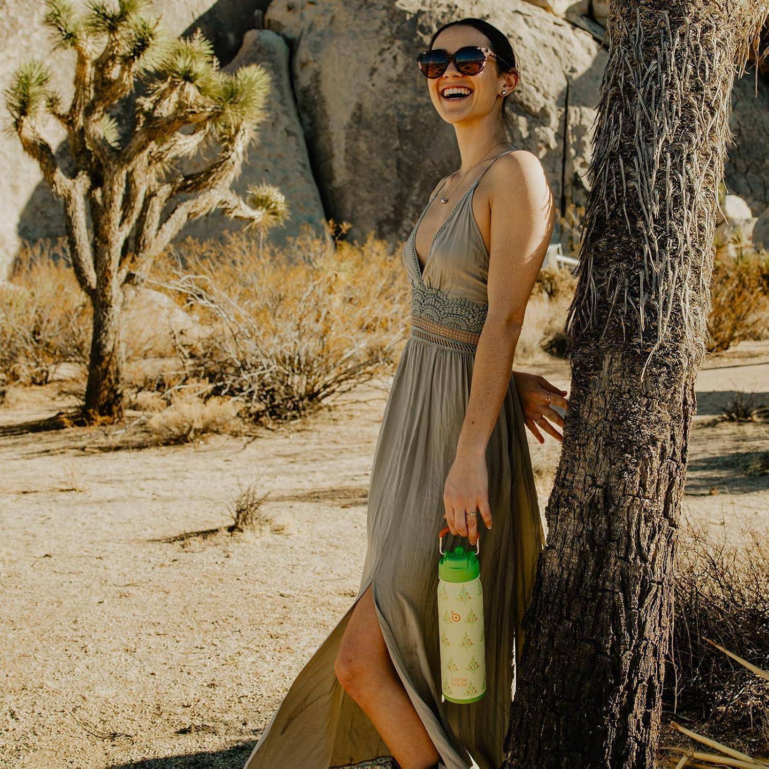 Eine Frau in einem weiten Kleid steht im Freien in der Nähe einer hohen Felsformation und eines Josua-Baums, hält eine grüne Wasserflasche und lächelt an einem sonnigen Tag.