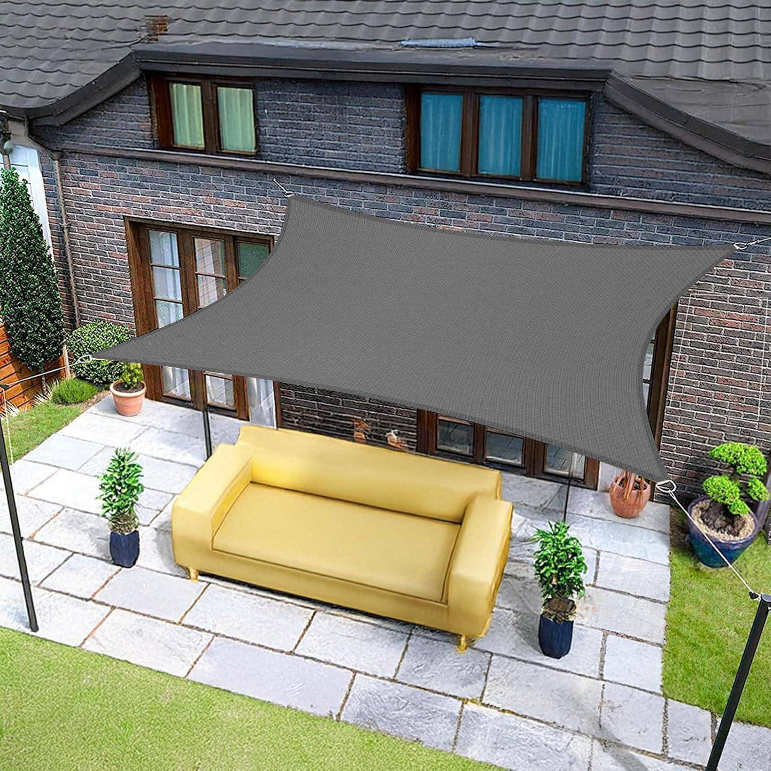 Eine Terrasse mit Steinfußboden zeigt eine gelbe Outdoor-Couch unter einem grauen Sonnensegel. Von Pflanzen umgeben, befindet sie sich neben einem Ziegelhaus.