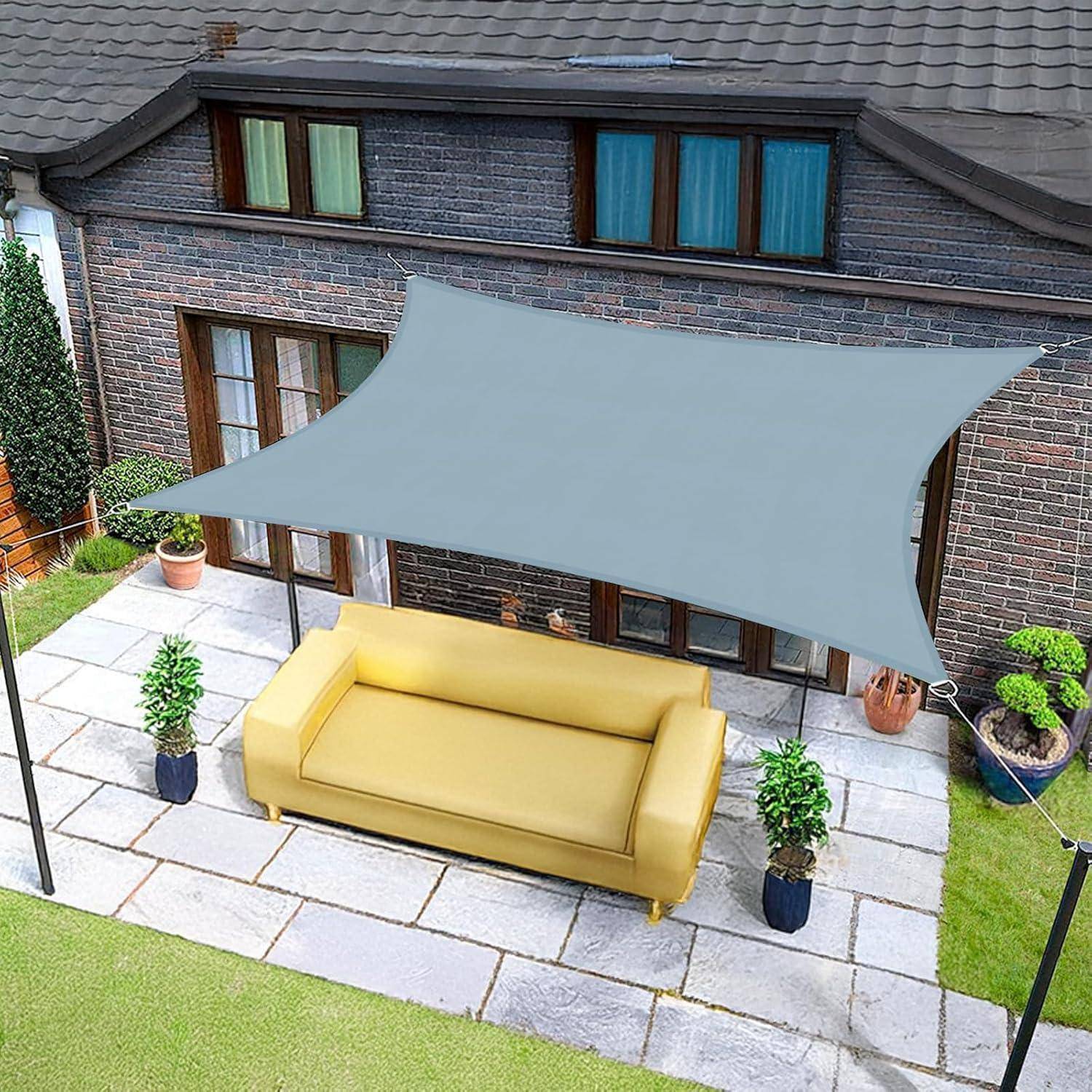 Eine gelbe Outdoor-Couch unter einem grauen Sonnensegel auf einer Terrasse neben einem Ziegelgebäude mit großen Fenstern und umgebender Vegetation.