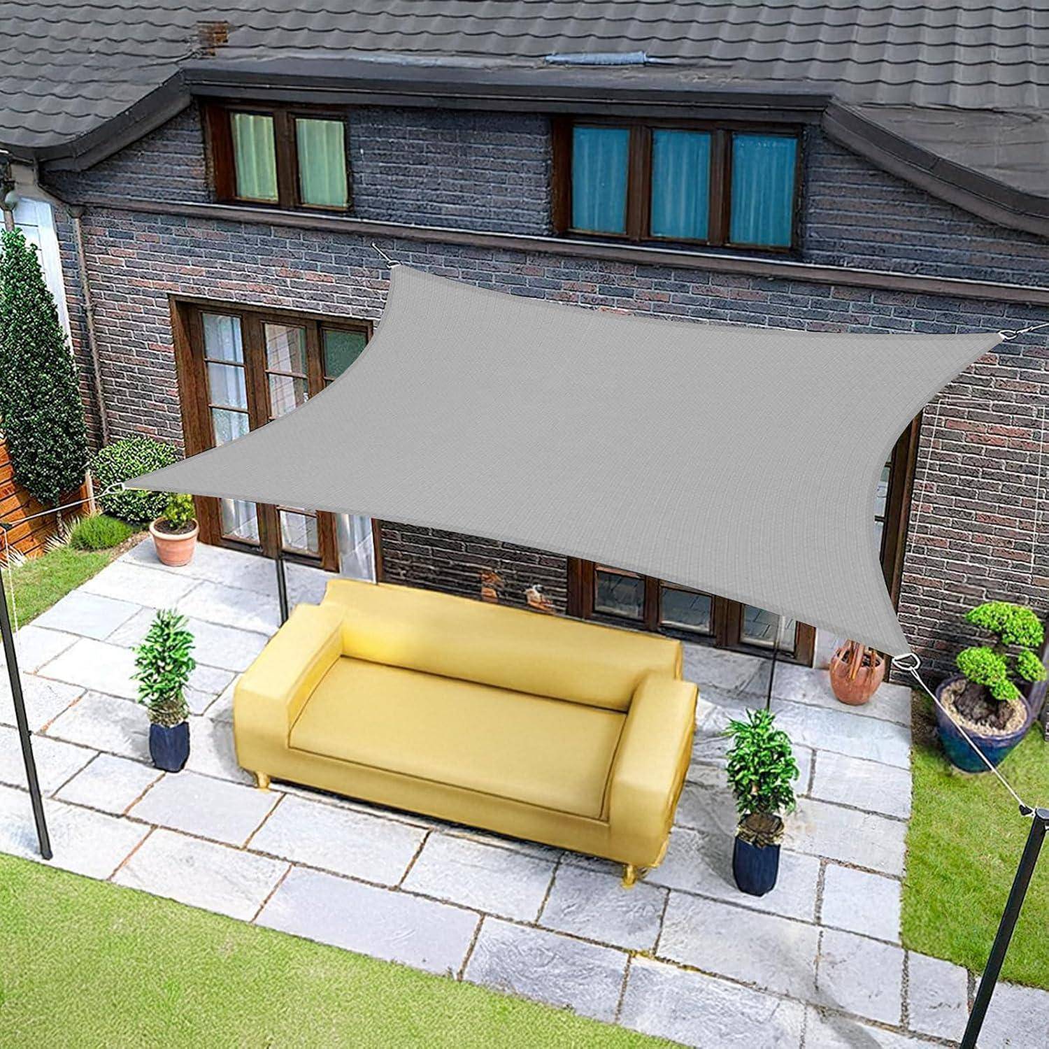 Eine Terrasse mit einem gelben Sofa unter einem grauen Sonnensegel. Der Bereich ist gepflastert, von Gras umgeben, mit Topfpflanzen und einem Ziegelgebäude.