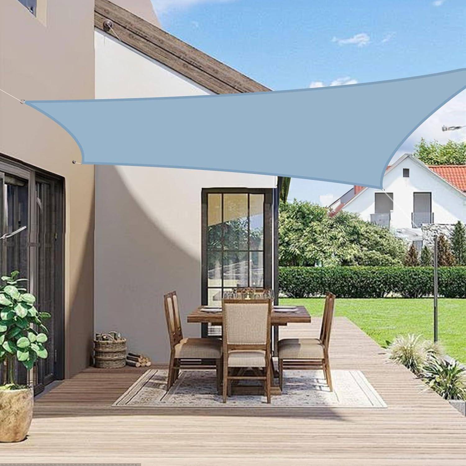 Outdoor-Terrasse mit einem Esstisch für sechs Personen unter einem blauen Sonnensegel. Der Bereich ist von Grünpflanzen umgeben und moderne Häuser befinden sich im Hintergrund.