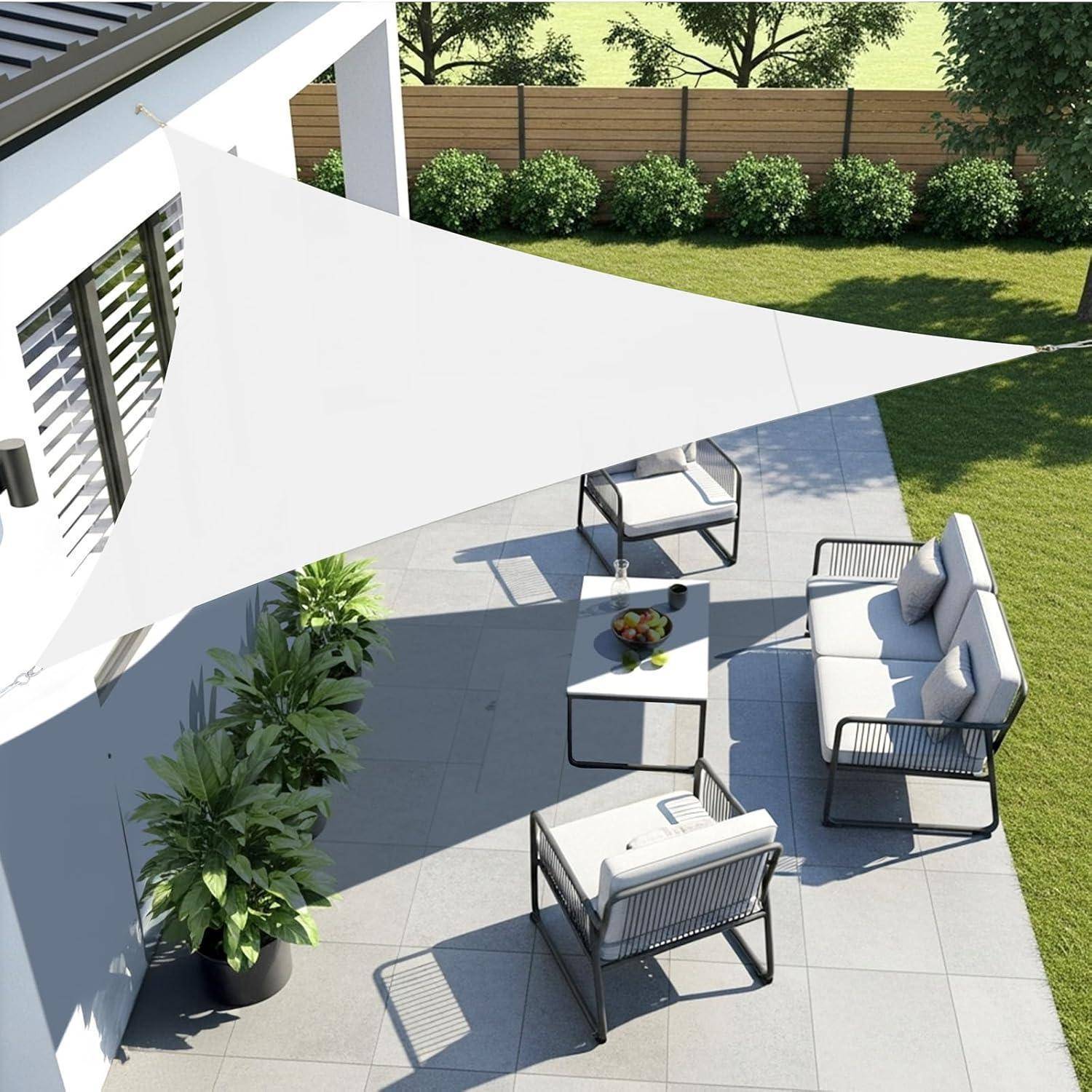 Eine moderne Terrasse mit einem weißen, dreieckigen Sonnensegel, einer Sitzgruppe mit grauen gepolsterten Stühlen und einem Tisch. Grüne Pflanzen und ein Holzzaun im Hintergrund.