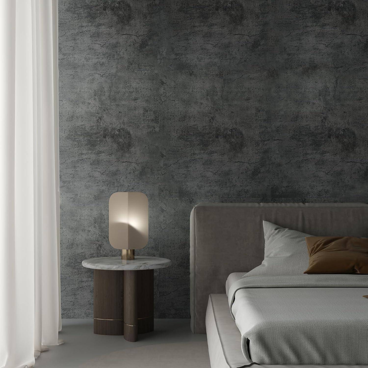 Ein minimalistisches Schlafzimmer mit einem grau gepolsterten Bett, einem braunen Kissen und einem kleinen runden Nachttisch mit einer Lampe. Weiße Vorhänge bedecken die linke Seite.