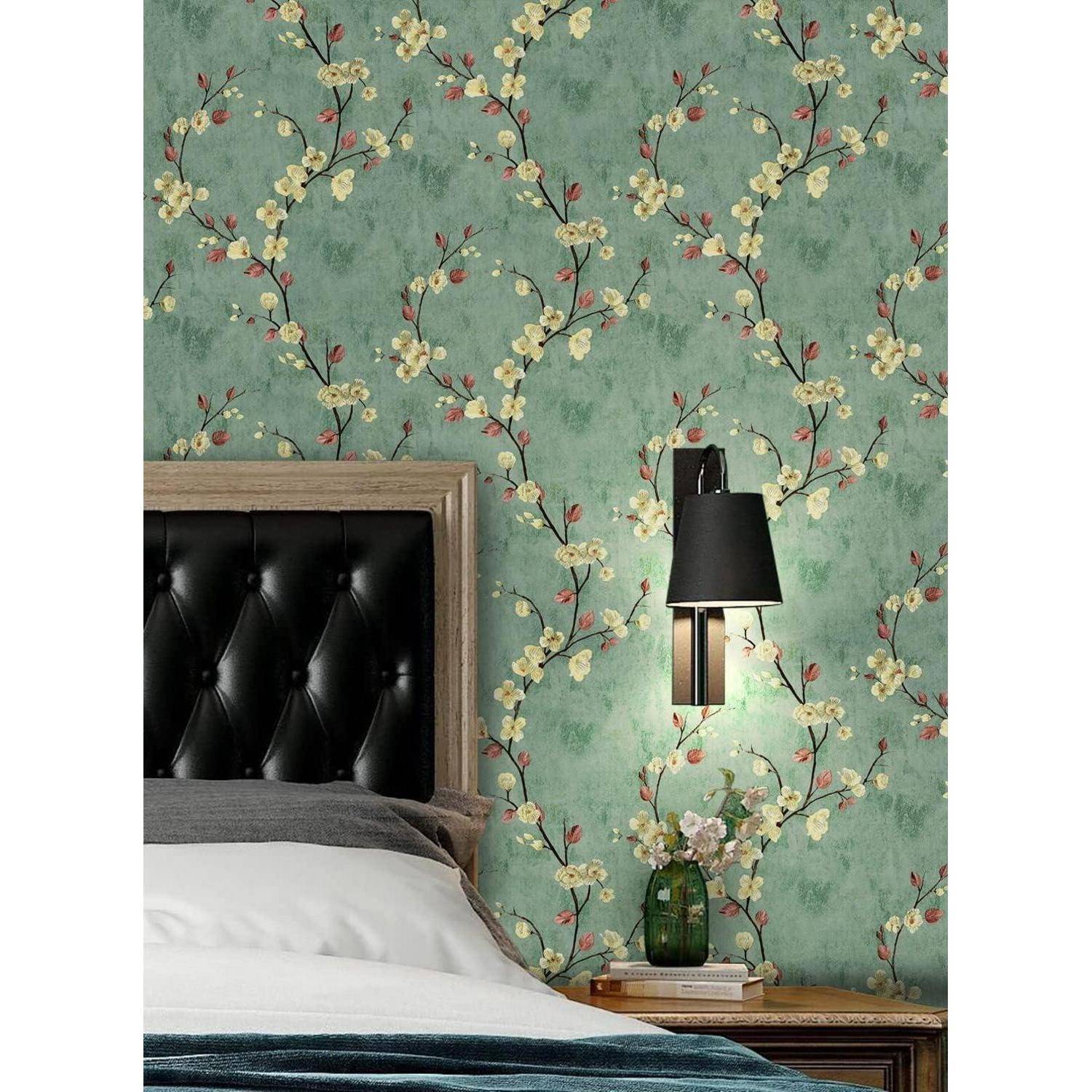 Ein Schlafzimmer zeigt einen schwarzen gepolsterten Kopfteil, eine blumenmusterartige Tapete und eine wandmontierte Lampe. Auf dem Nachttisch stehen eine kleine Pflanze und ein Buch.