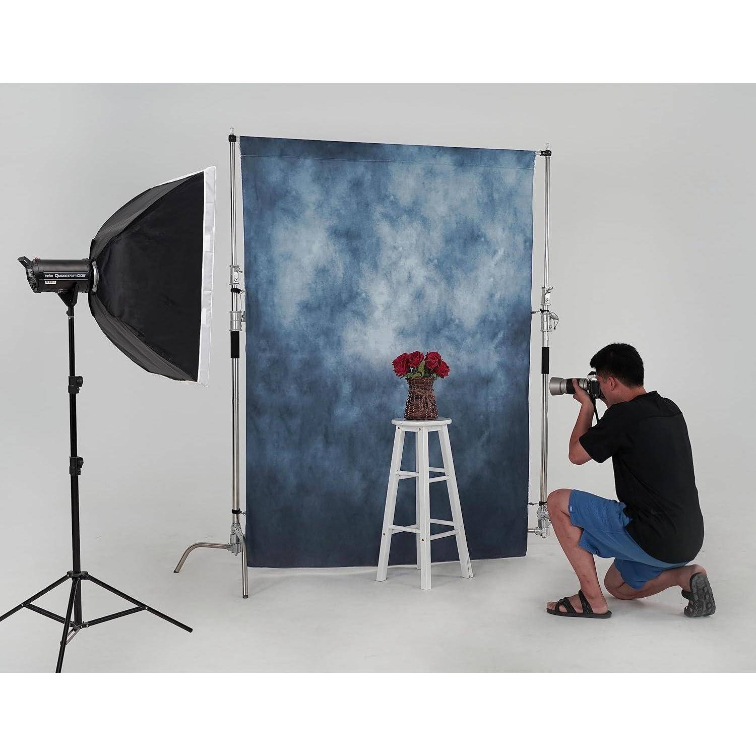 Ein Fotograf in kurzen Hosen kniet und macht ein Foto eines Hockers mit einer Vase roter Rosen vor einem blauen Hintergrund in einem Fotostudio-Ambiente.