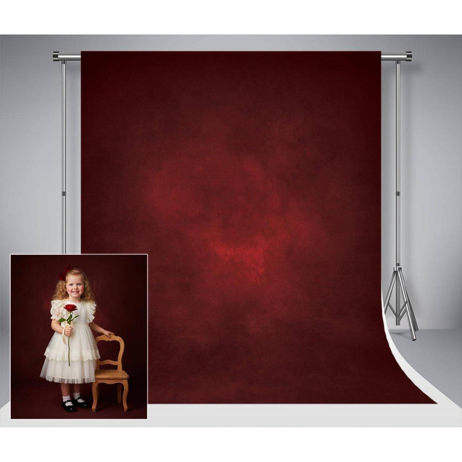 Ein junges Kind in einem weißen Kleid, das eine rote Rose hält, steht neben einem Holzstuhl vor einem großen, dunklen roten Studiohintergrund.