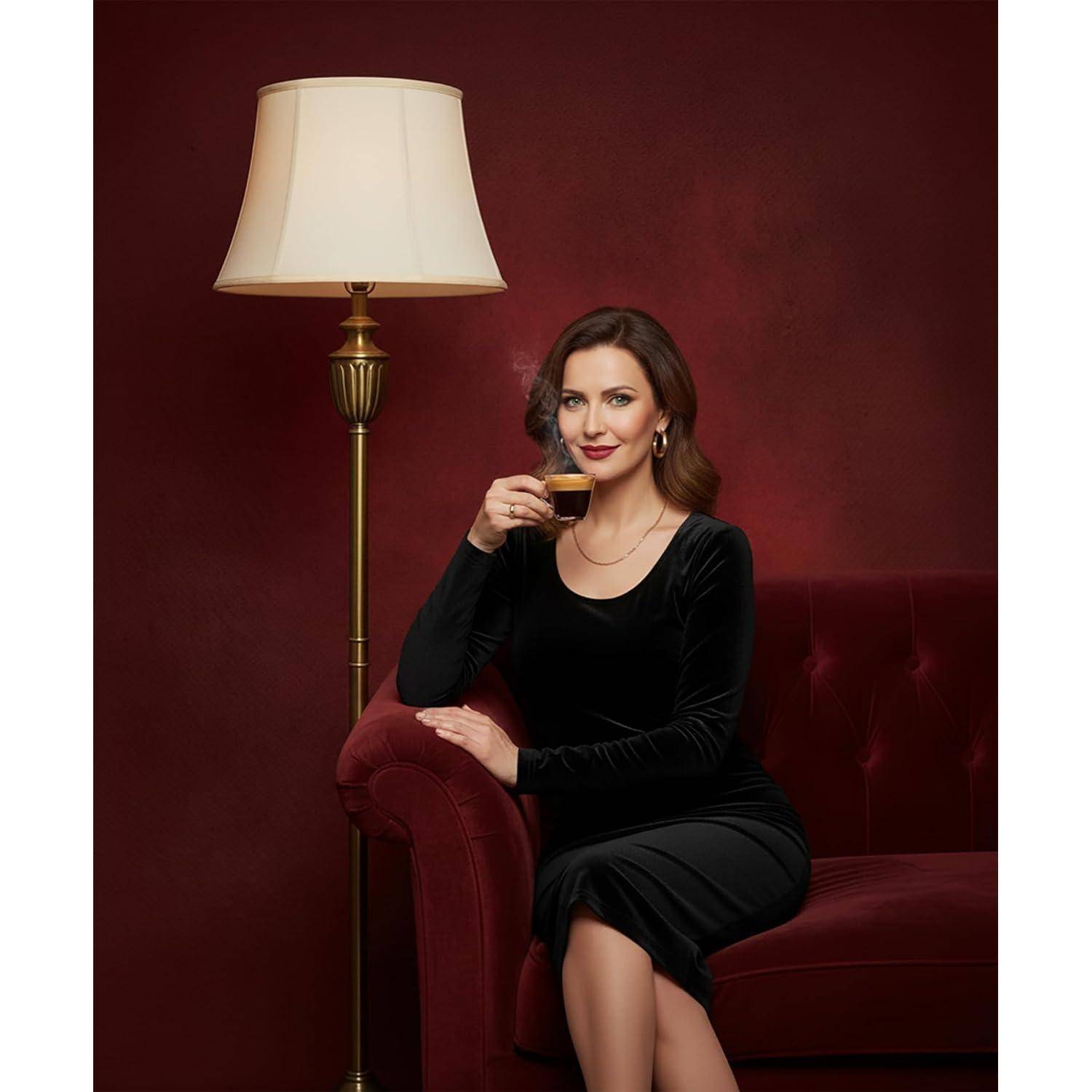 Eine Frau in einem schwarzen Kleid sitzt auf einem roten Sofa und genießt Kaffee neben einer hohen Lampe vor einem tiefroten Hintergrund.