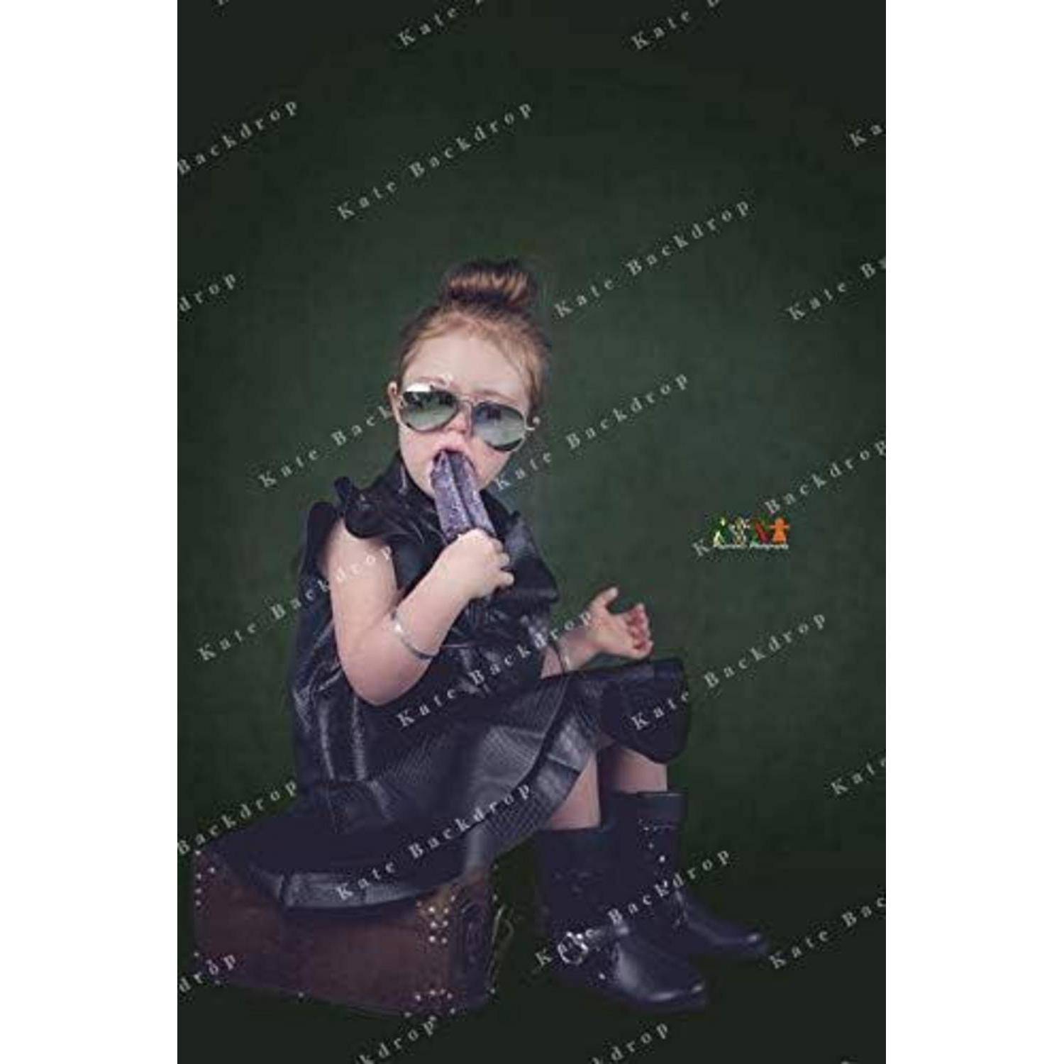 Ein junges Kind sitzt auf einem Koffer, trägt ein schwarzes Kleid und Sonnenbrille und hält ein Stück violetten Stoff vor einem dunkelgrünen Hintergrund.