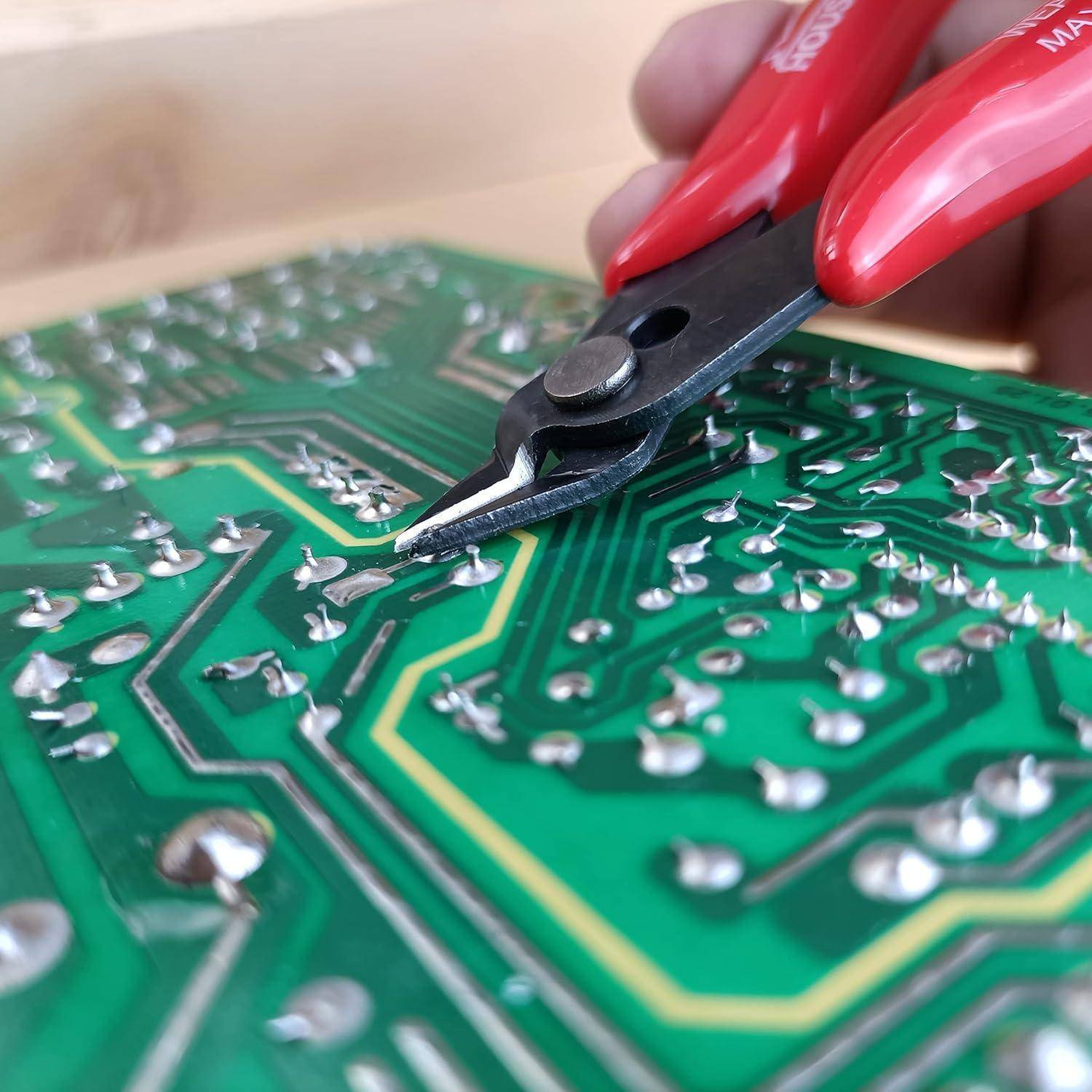 Nahaufnahme einer Hand, die eine Zange mit roten Griffen verwendet, um an einer grünen Leiterplatte mit silbernen Komponenten zu arbeiten, was auf eine Elektronik-Reparatur hinweist.