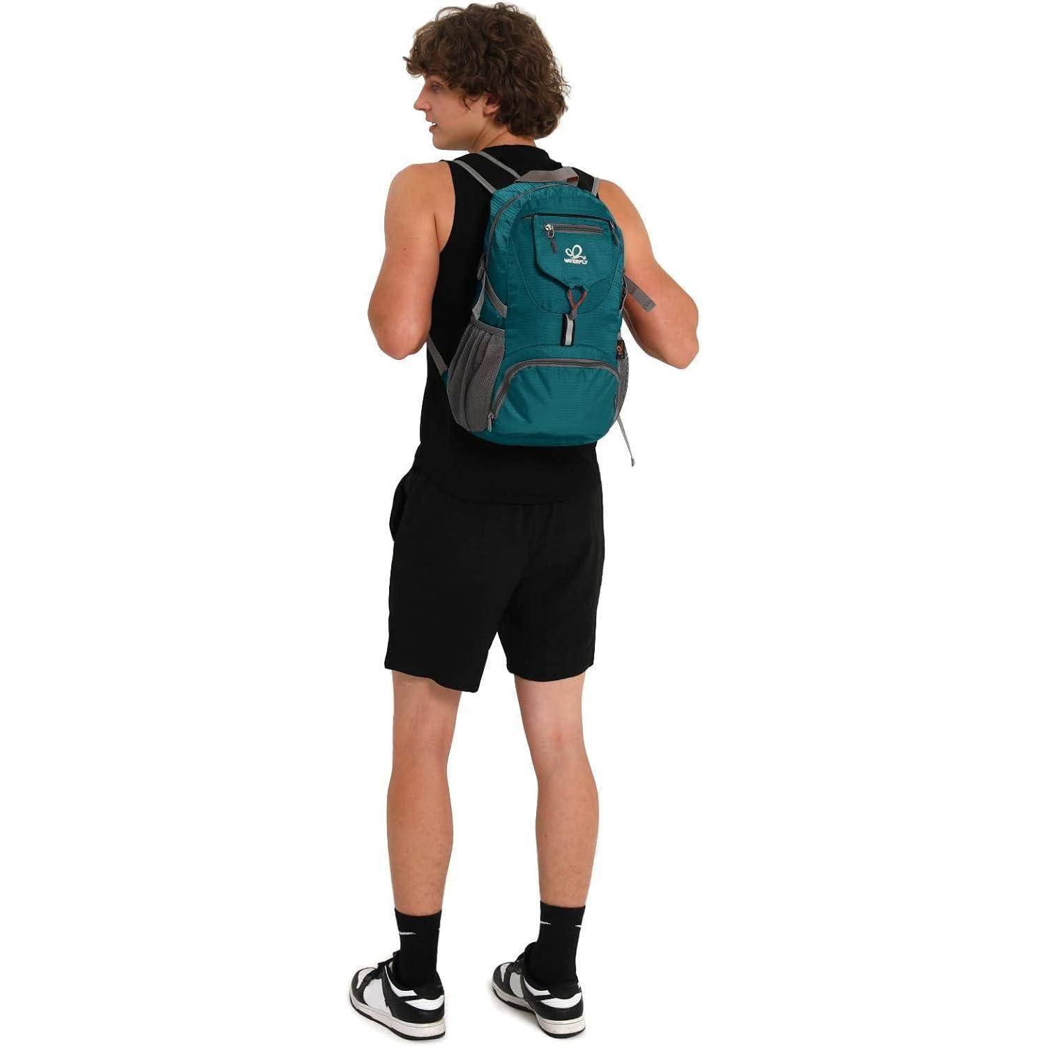 Eine Person mit lockigem Haar, die ein schwarzes Trägertop, Shorts und Turnschuhe trägt, steht mit einem petrolblauen Rucksack in einem Innenraum.