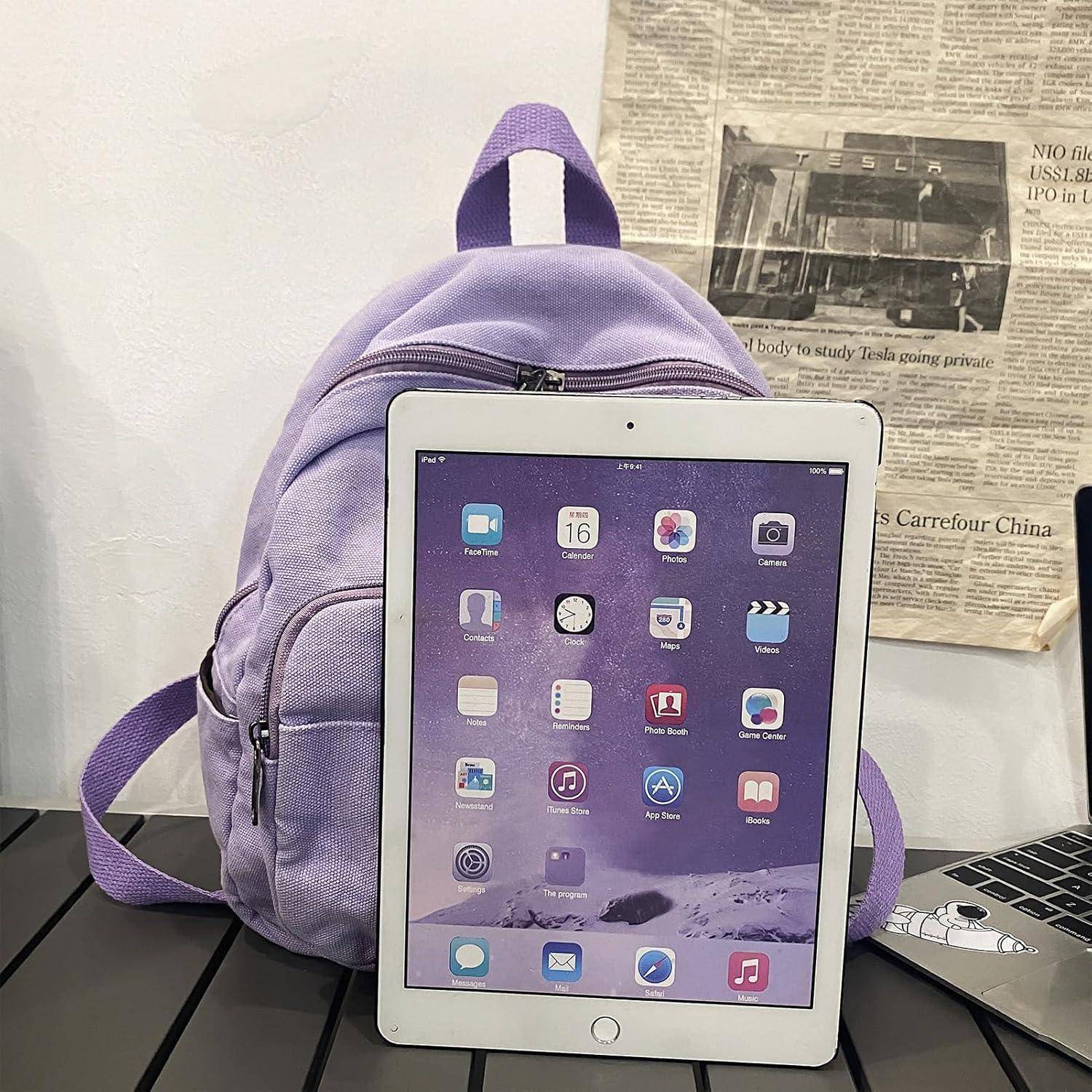 Ein Lavendelrucksack liegt auf einem schwarzen Tisch neben einem iPad, auf dessen Startbildschirm App-Symbole zu sehen sind. Eine Zeitung ist an der Wand befestigt.