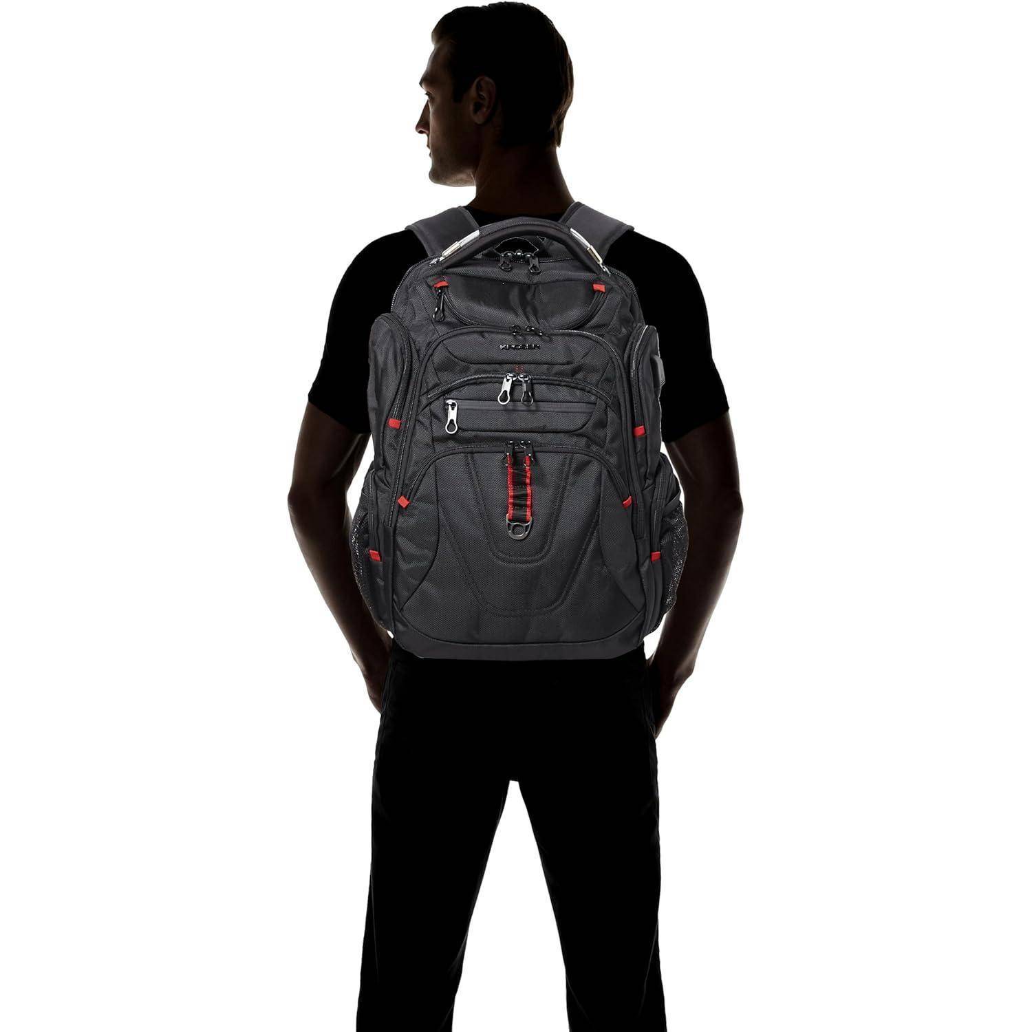 Eine Person in einem schwarzen T-Shirt steht mit dem Rücken zur Kamera und trägt einen großen Rucksack in Grau und Rot mit mehreren Taschen und Riemen.