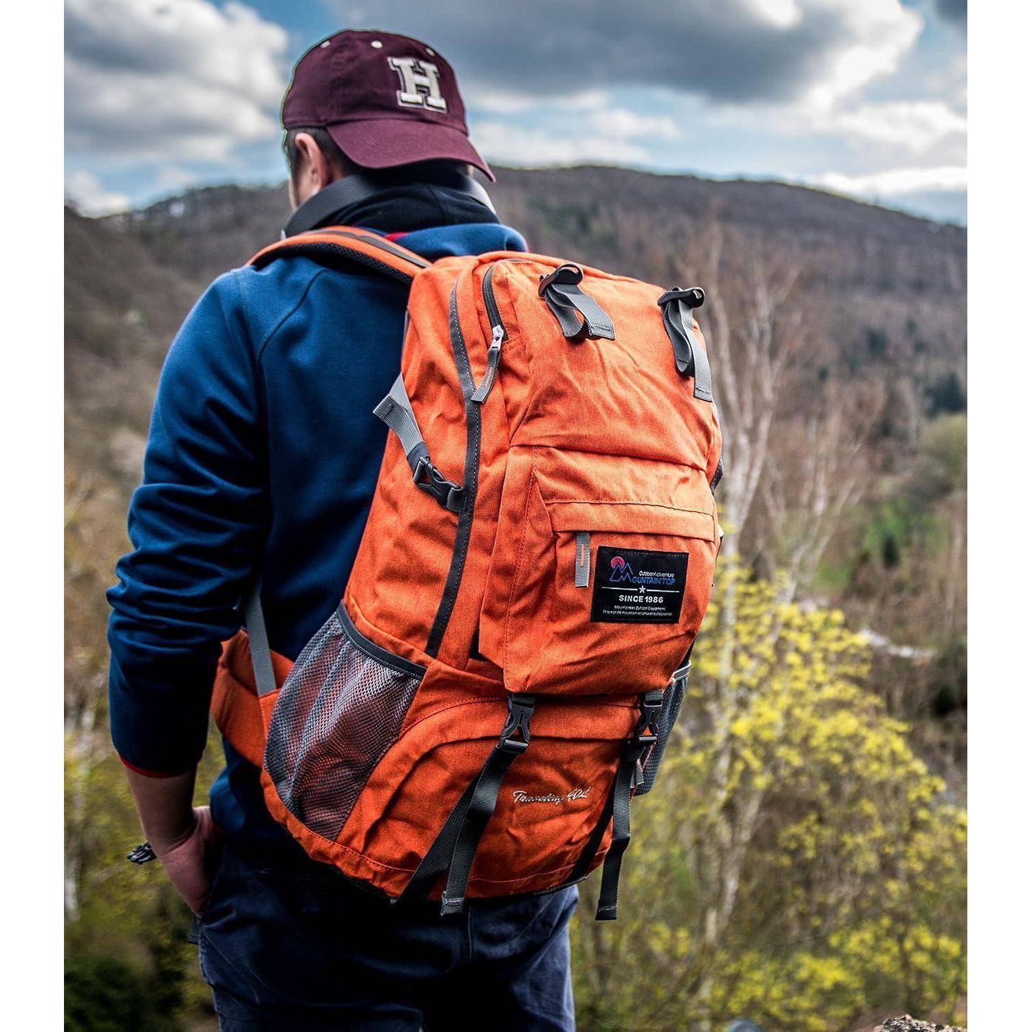 Eine Person trägt eine marronrote Kappe und einen blauen Hoodie und hat einen orangefarbenen Rucksack, während sie auf einem bewaldeten Hügel mit einem bergigen Hintergrund steht.
