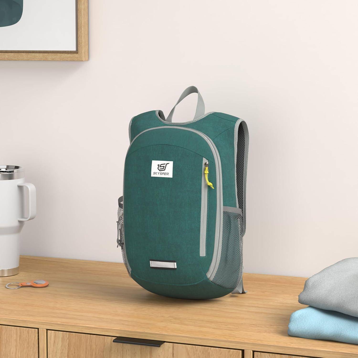 Ein grüner Rucksack mit einem gelben Reißverschluss liegt auf einem Holzschreibtisch. Eine Tasse, ein Schlüsselanhänger und ein gefaltetes Handtuch befinden sich in der Nähe. Ein gerahmtes Bild hängt an der Wand.