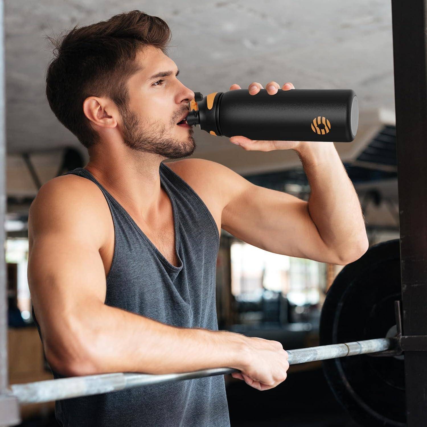 Ein Mann in einem ärmellosen Top trinkt aus einer schwarzen Wasserflasche in einem Fitnessstudio, in der Nähe von Trainingsgeräten, und wirkt konzentriert und nach dem Training.