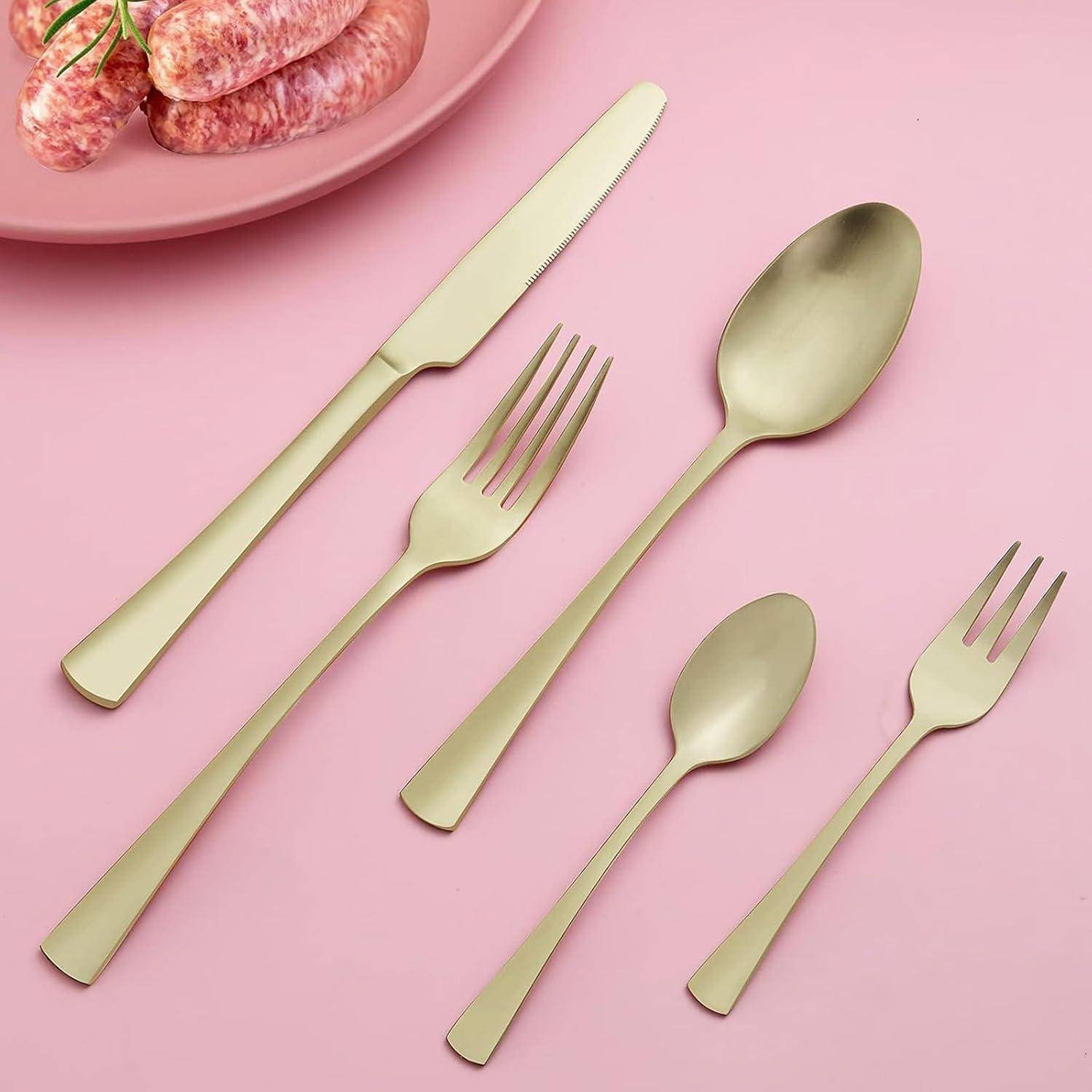 Goldfarben gehaltenes Besteckset mit Messern, Löffeln und Gabeln, das auf einem pinken Hintergrund neben einem Teller mit Würstchen präsentiert wird.