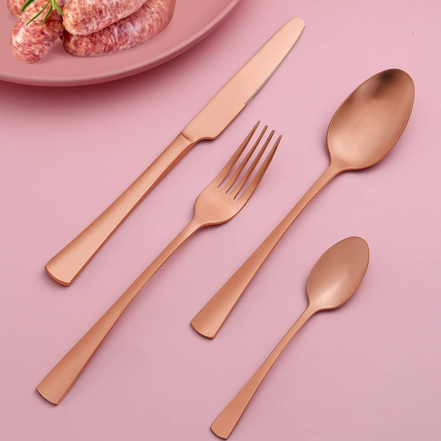 Ein Set Roségold-Besteck, bestehend aus einem Messer, einer Gabel, einem Teelöffel und einem Esslöffel, elegant auf einer rosa Oberfläche angeordnet, mit einem Teller mit Würstchen im Hintergrund.