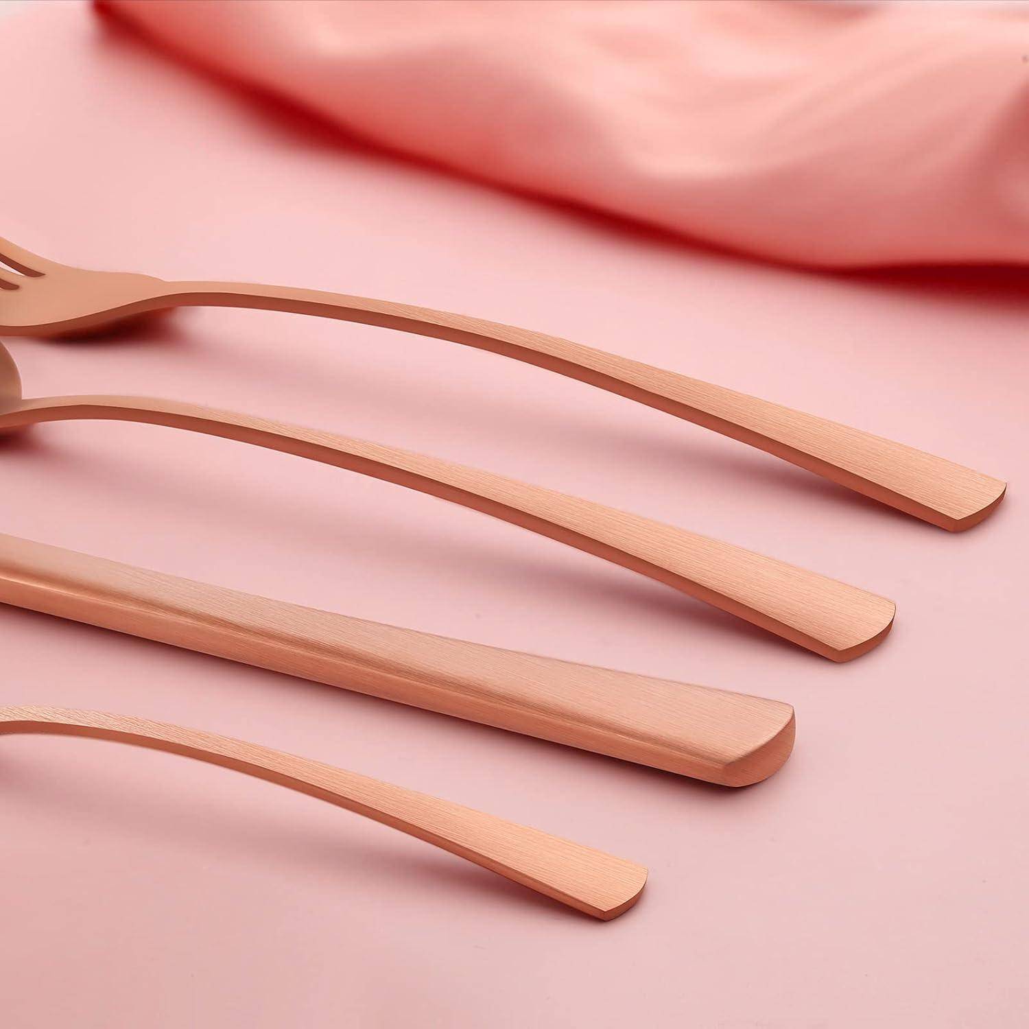 Eine Nahaufnahme von elegantem Roségold-Besteck, einschließlich einer Gabel, Messern und einem Löffel, diagonal auf einer weichen rosa Oberfläche angeordnet.