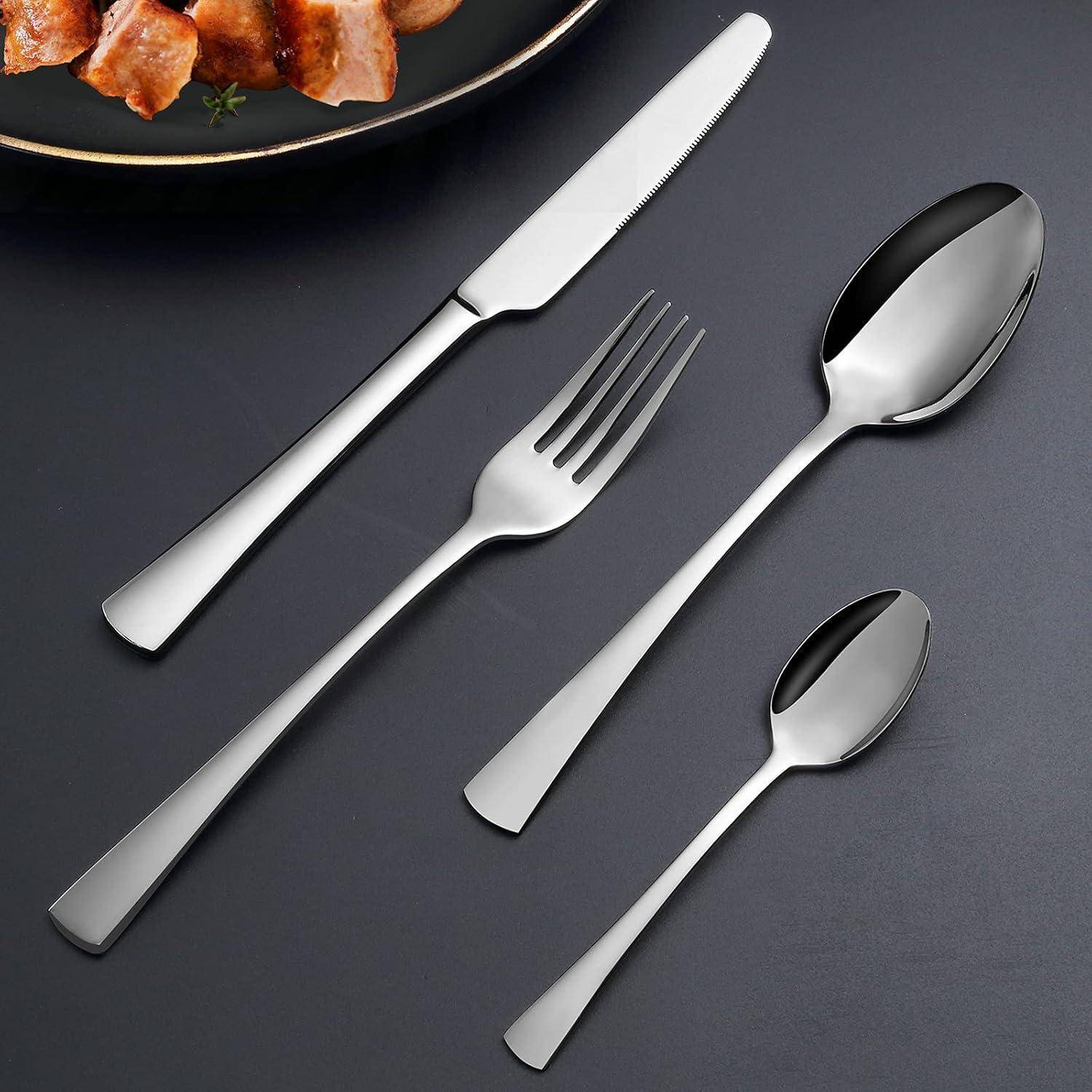 Ein Set Besteck aus Edelstahl, bestehend aus einem Messer, einer Gabel, einem Esslöffel und einem Teelöffel, angeordnet auf einer dunklen Oberfläche neben einem Teller mit gebratenem Fleisch.