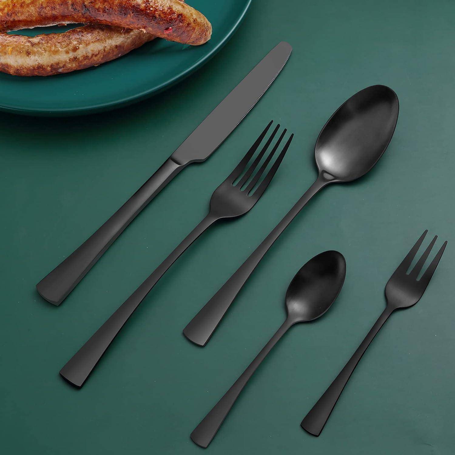 Ein Set matter schwarzer Bestecke, bestehend aus einem Messer, einer Gabel, einer Dessertgabel, einem Löffel und einem Teelöffel, die neben einem Teller mit zwei Würstchen angeordnet sind.