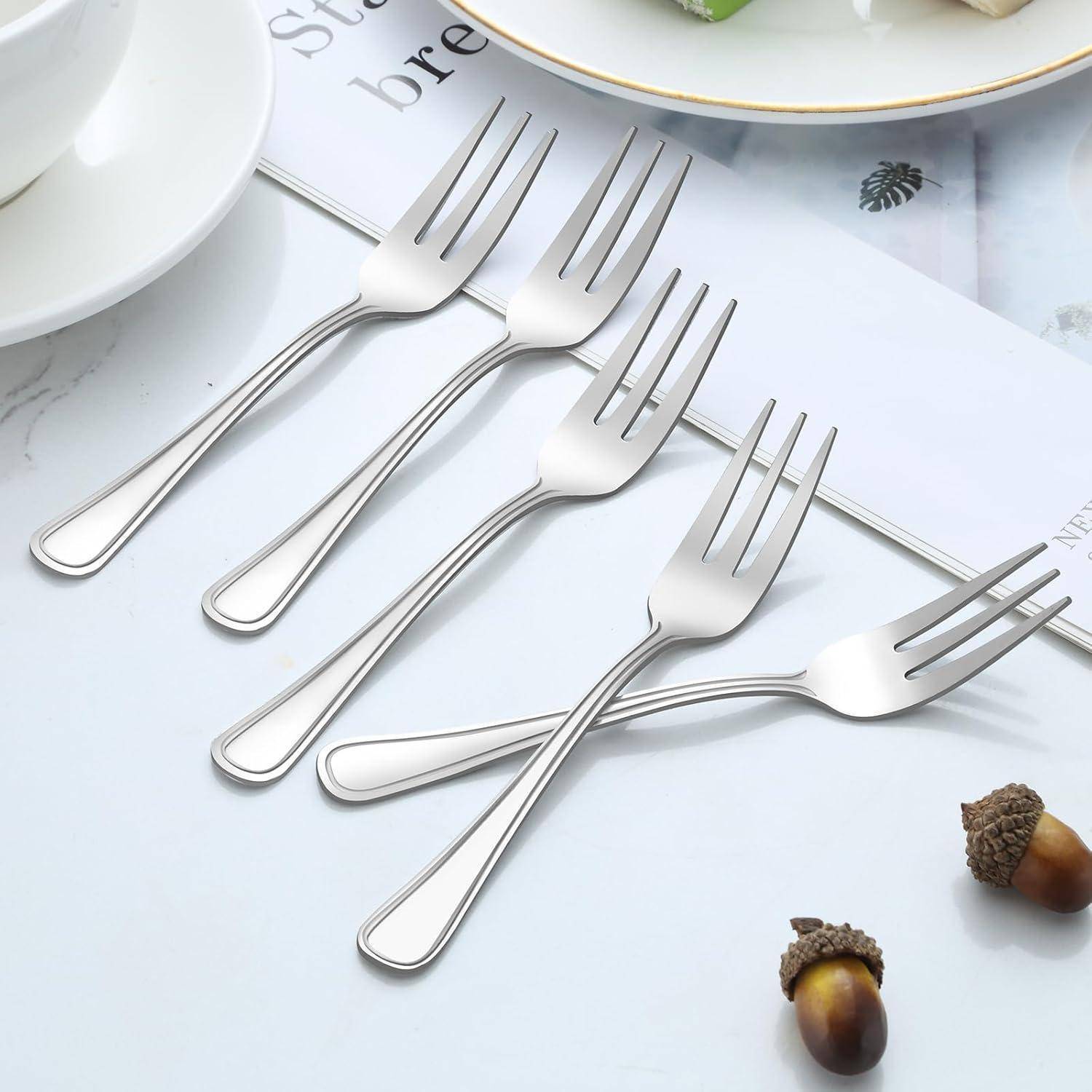 Ein Satz aus sechs silbernen Gabeln, elegant auf einer weißen Oberfläche angeordnet, mit Eicheln in der Nähe, was eine minimalistische Tischgestaltung andeutet.