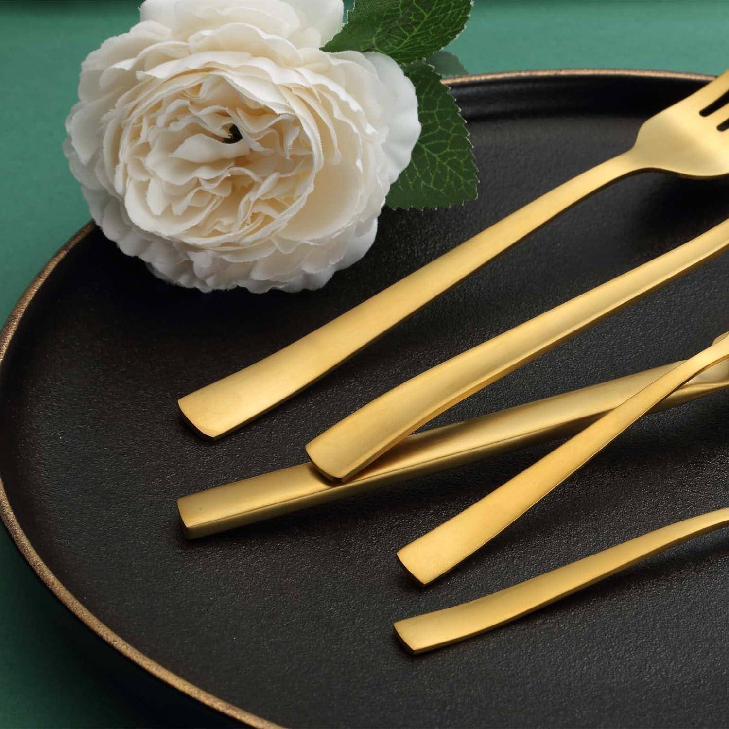 Eine weiße Rose und elegantes goldenes Besteck, bestehend aus einem Messer, einer Gabel und einem Löffel, ruhen auf einem dunklen Teller vor einem grünen Hintergrund.