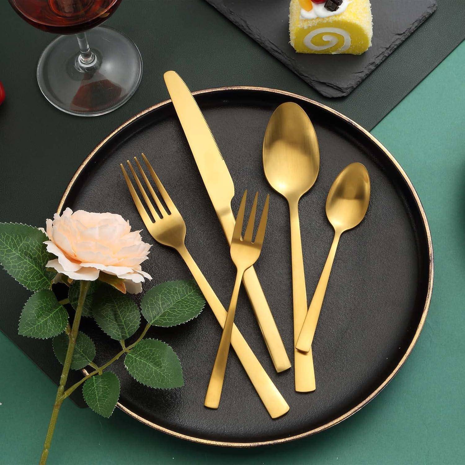 Goldenes Besteck-Set auf einem schwarzen Teller mit einem Messer, einer Gabel, einem Löffel und einer Dessertgabel. In der Nähe eine Rose und ein Dessertgebäck, dazu ein Glas Rotwein.