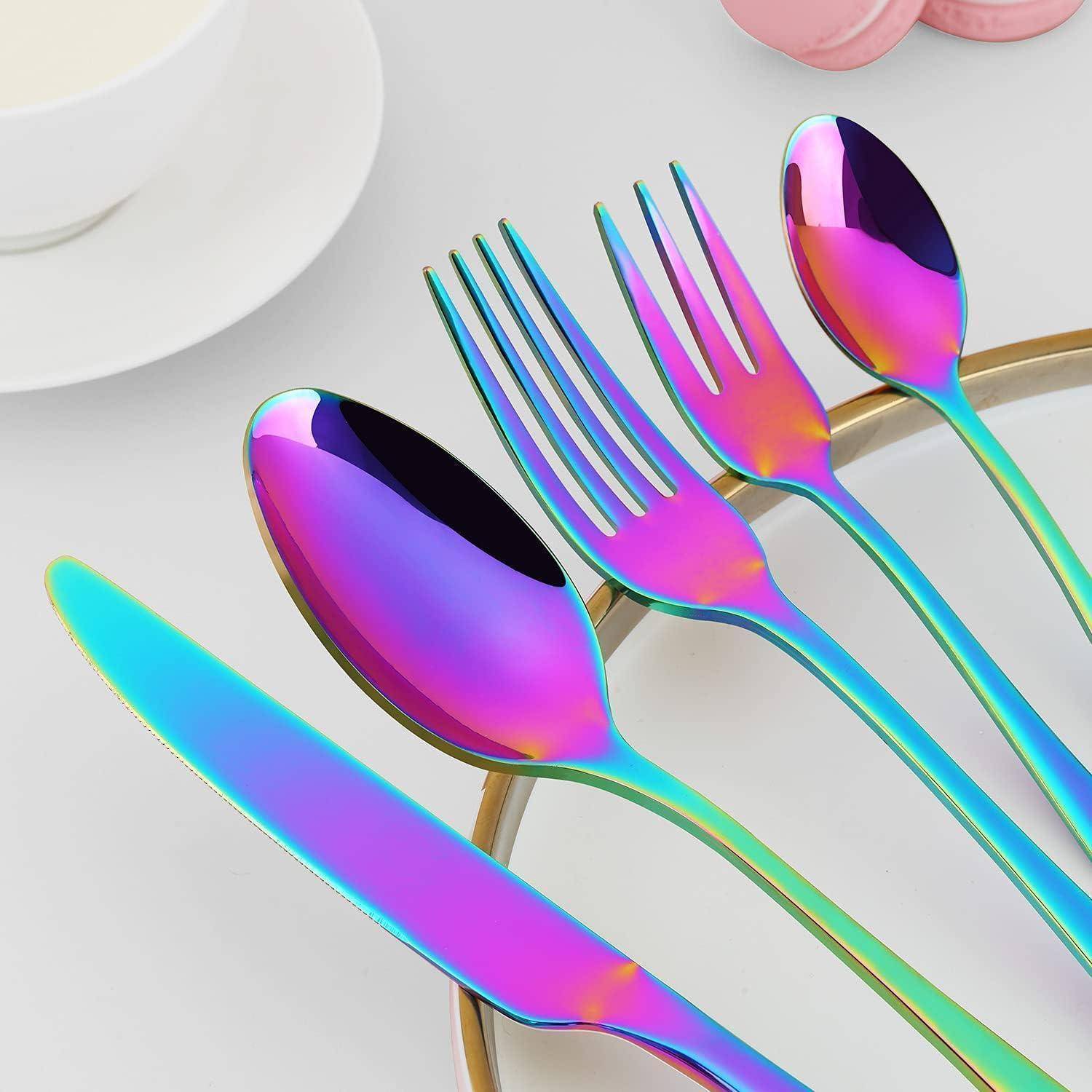 Schillerndes Besteckset mit einem Löffel, einer Gabel und einem Messer, das Regenbogenfarben auf einer hellgrauen Oberfläche in der Nähe einer weißen Tasse zeigt.