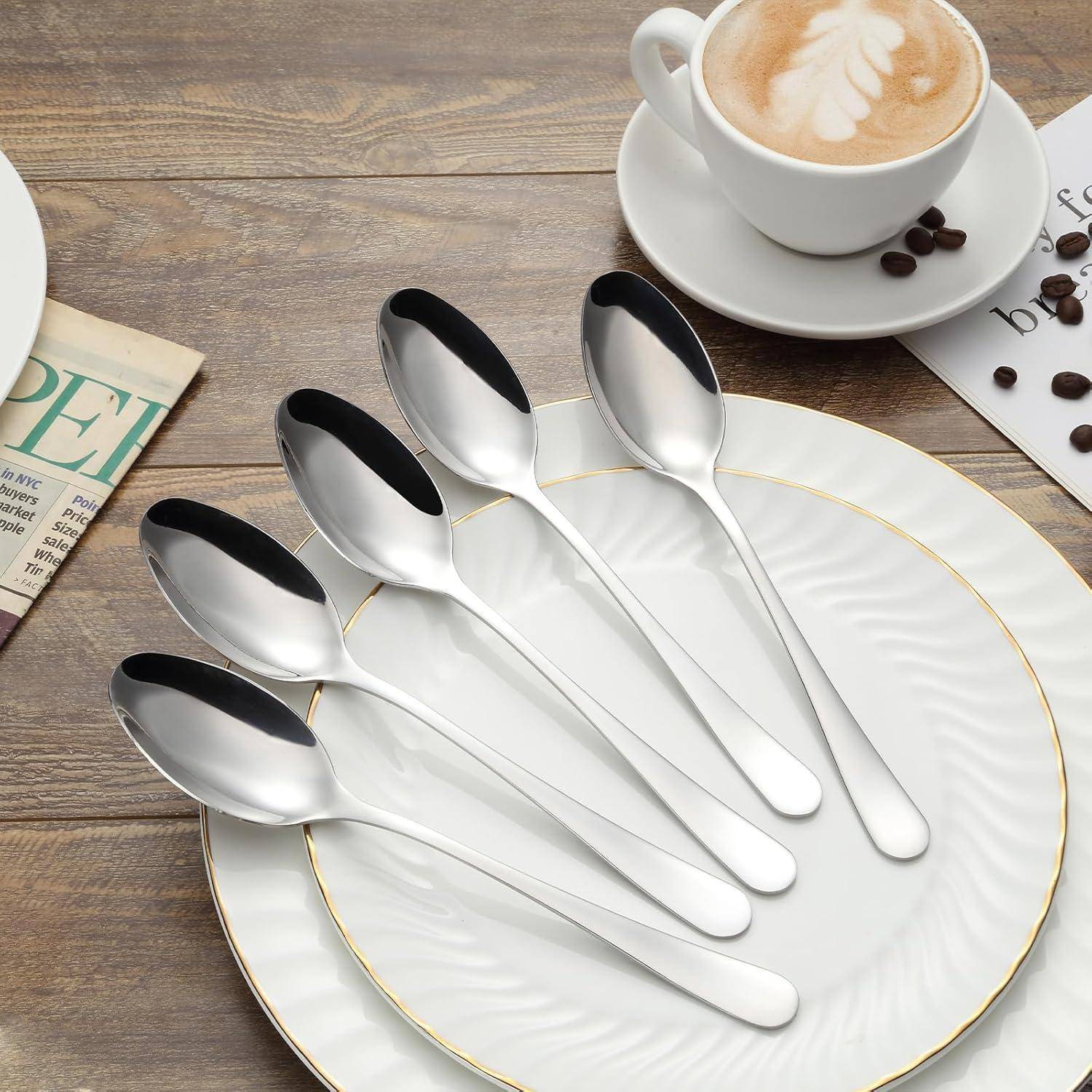 Sechs silberne Löffel, die in einer Fächerform auf einem weißen Teller neben einer Tasse Kaffee auf einem Holztisch angeordnet sind.