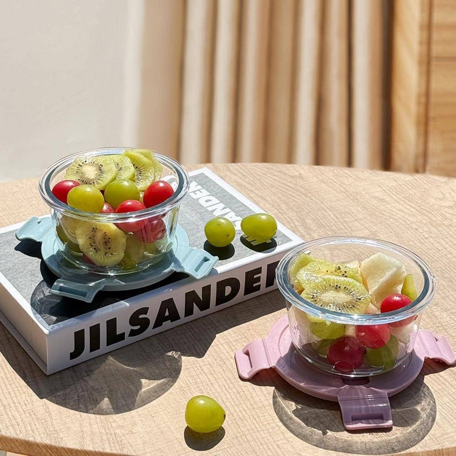 Zwei Glasschalen mit geschnittenen Früchten, darunter Kiwi und Trauben, stehen auf einem Holztisch bei Sonnenlicht, daneben ein Buch mit dem Titel „Jil Sander
