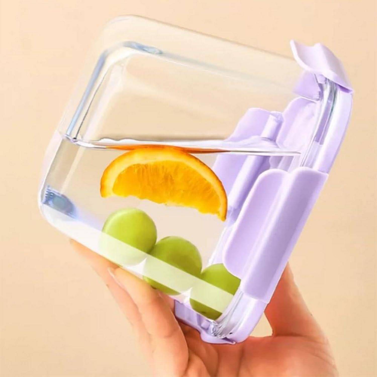 Eine Hand hält eine durchsichtige Box, gefüllt mit Wasser und drei grünen Trauben, die von einem Orangenschnitz gekrönt wird. Die Box hat einen hellvioletten Deckel.