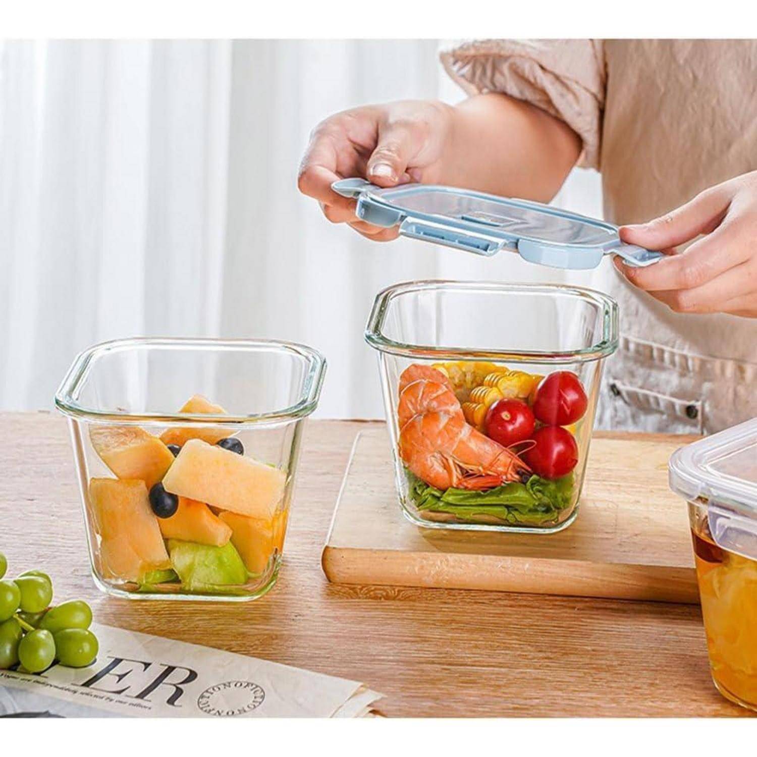 Eine Person verwendet blaue Deckel, um Lebensmittelbehälter zu verschließen, die mit Obst, Gemüse und Salat gefüllt sind. Trauben sind in der Nähe auf einem Tisch sichtbar.