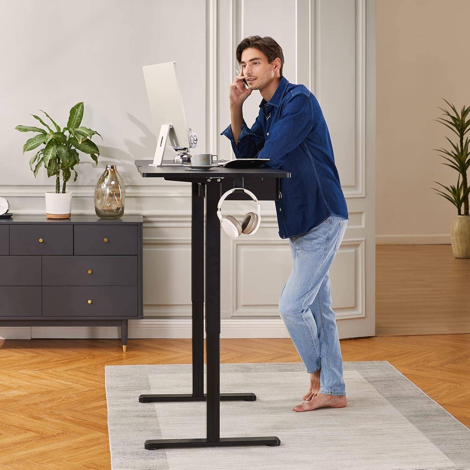Eine Person arbeitet an einem Stehschreibtisch in einem hellen Raum mit Pflanzen, trägt lässige Kleidung und blickt auf einen Computerbildschirm.