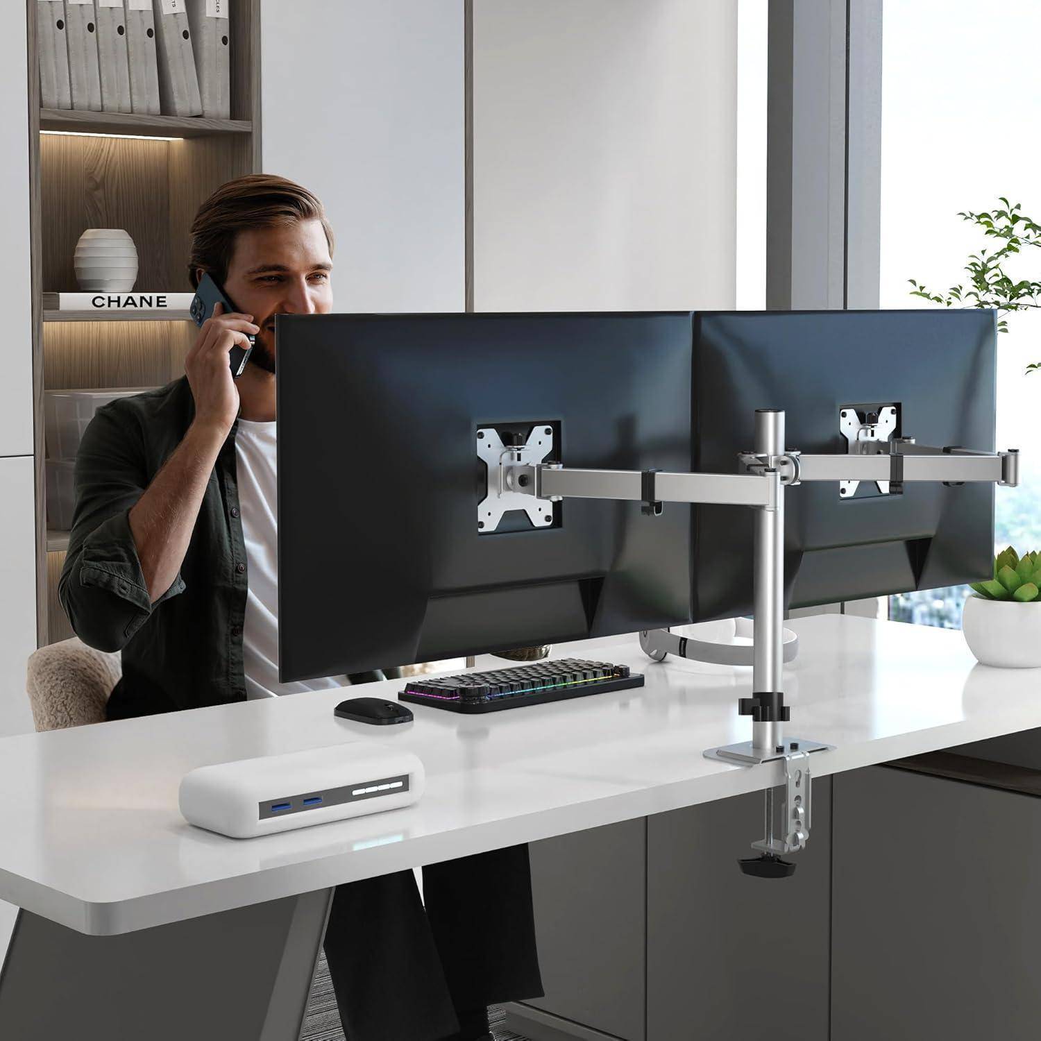 Eine Person sitzt an einem Schreibtisch und telefoniert, während sie auf zwei Monitore auf einem verstellbaren Ständer blickt. Die Umgebung ist ein modernes Büro.