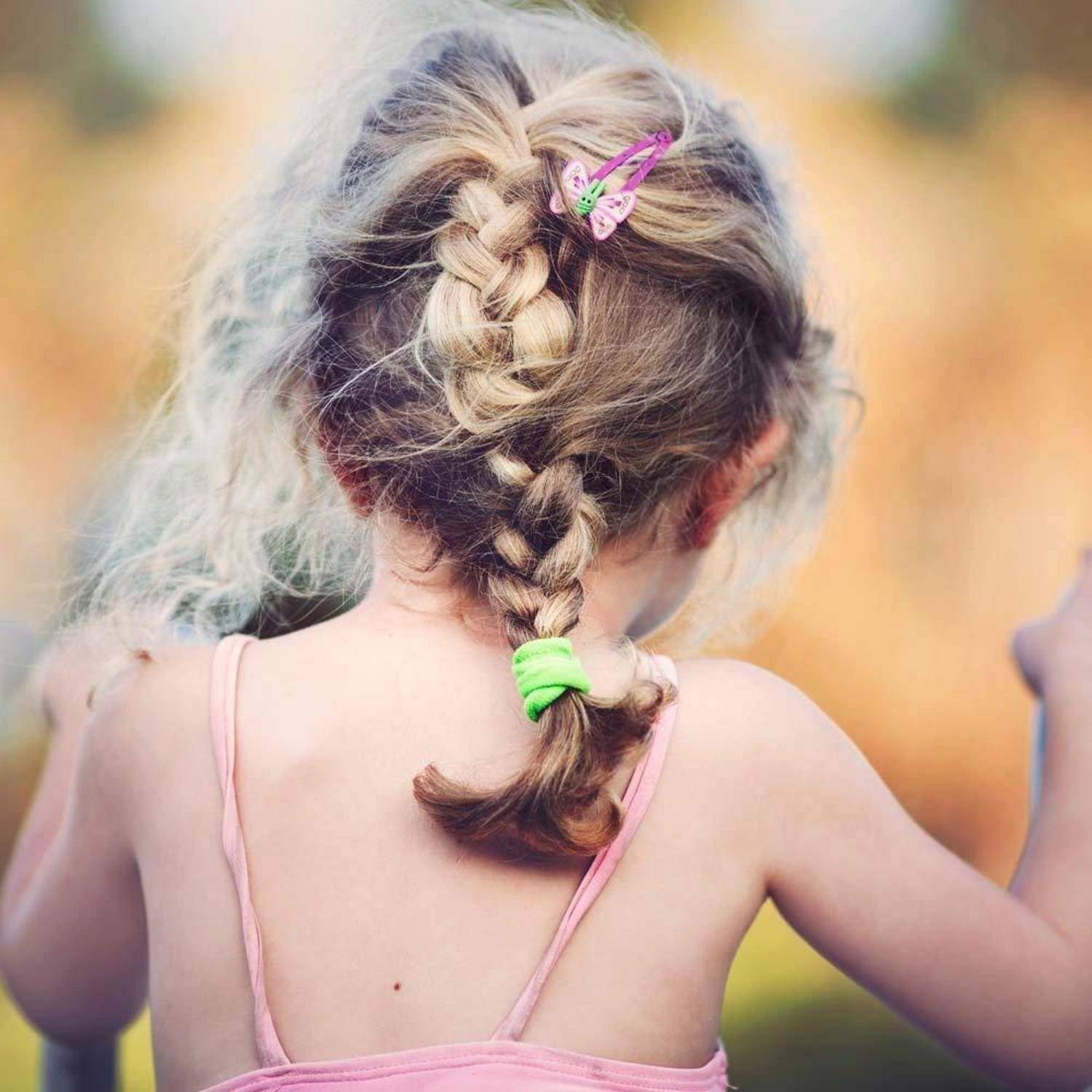 Ein junges Mädchen mit blonden Haaren, zu einem Zopf geflochten, geschmückt mit einer Schmetterlingsklammer und einem grünen Haargummi, von hinten in einer sonnendurchfluteten Umgebung.