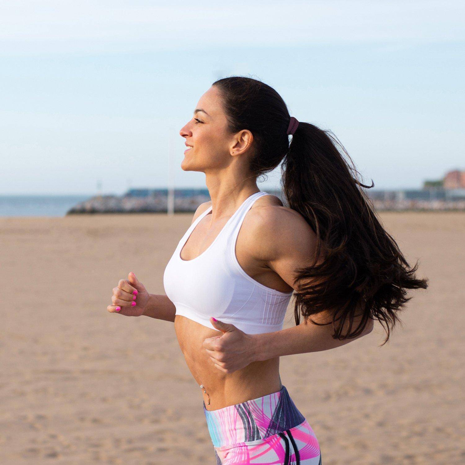 Eine Frau joggt an einem Strand, trägt ein weißes Trägertop und rosa Shorts, mit einem langen Pferdeschwanz, konzentriert darauf, direkt am Ozean Sport zu treiben.