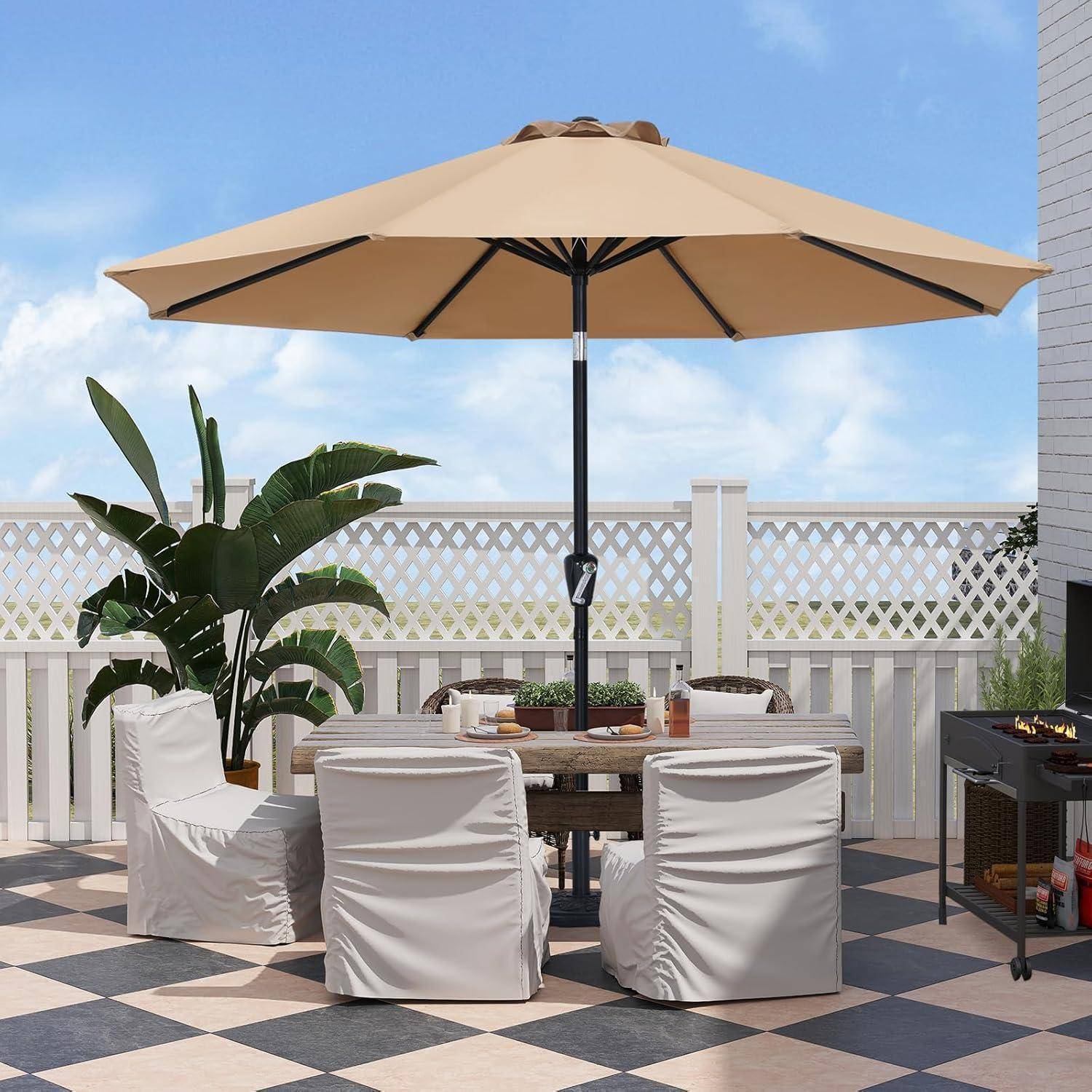Eine Terrasse mit einem großen beigen Sonnenschirm beschattet einen Tisch und sechs weiße Stühle auf einem karierten Boden. Eine Topfpflanze und ein weißer Zaun befinden sich im Hintergrund.